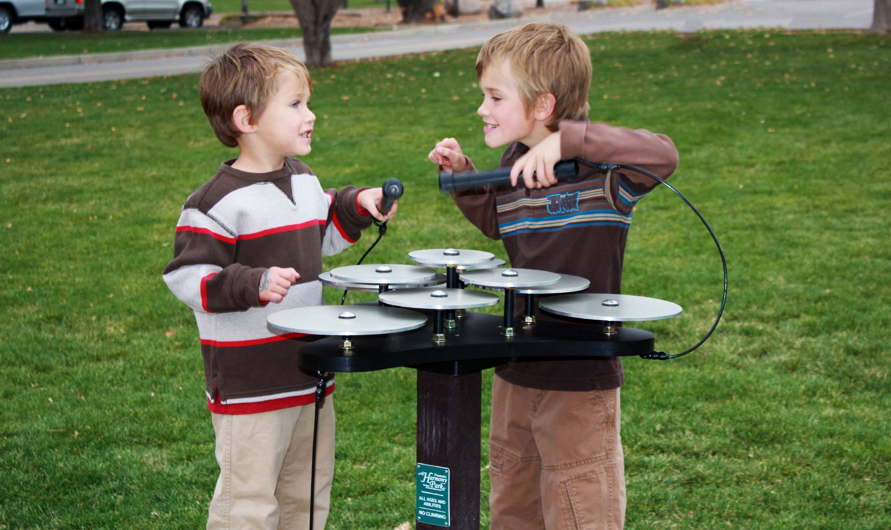 Two children playing on a black outdoor musical instrument with ten silver anodized aluminum cymbal discs and attached mallets