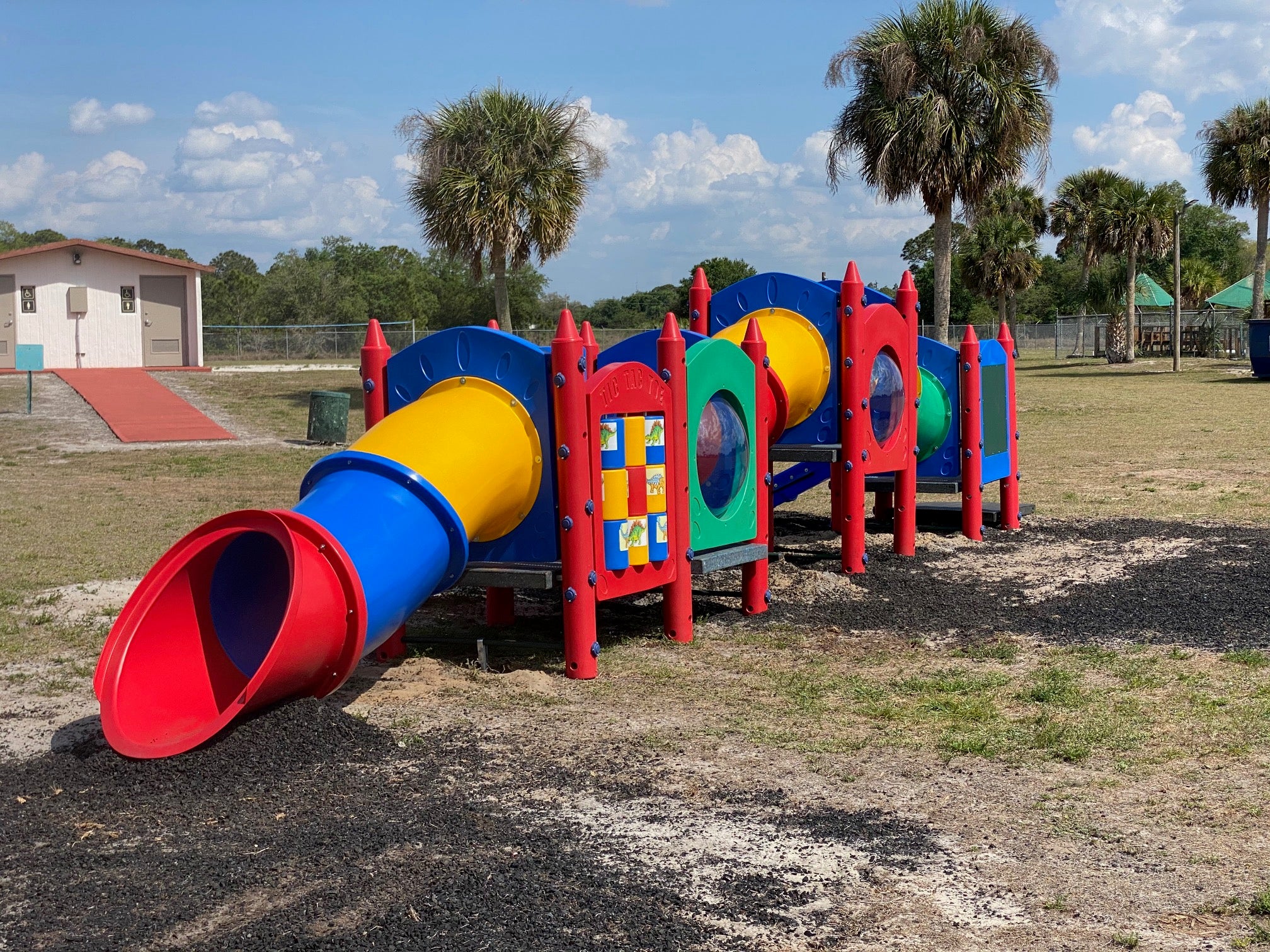 Colorful low-profile playground with crayon-themed towers, interactive panels, crawl tunnel, and red tube slide on grass with palm trees.