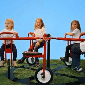 Children riding a red merry-go-round cycle with seats and pedals on green grass under clear blue sky
