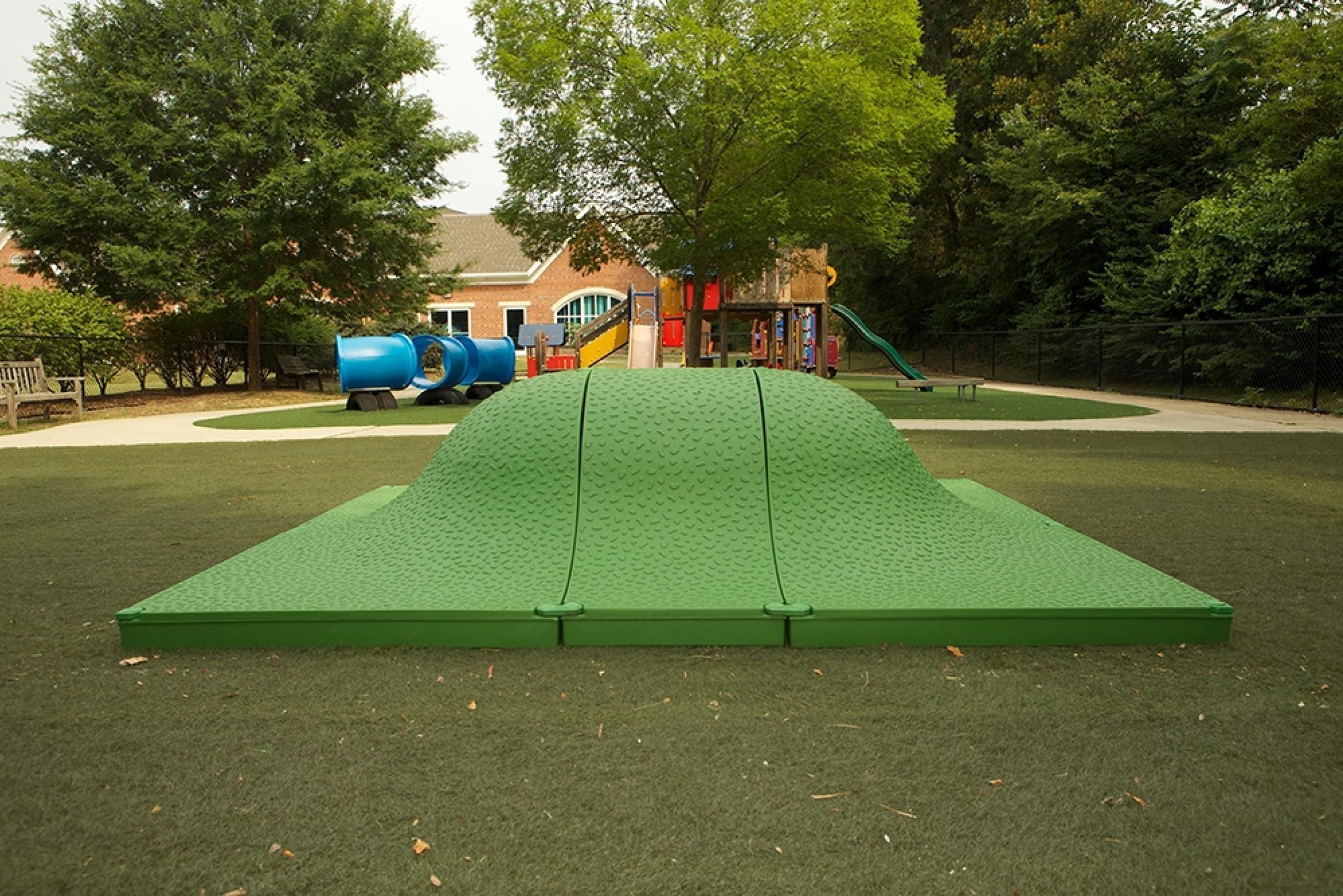 Green textured play mound with rounded peak on artificial turf, surrounded by climbing and sliding playground equipment