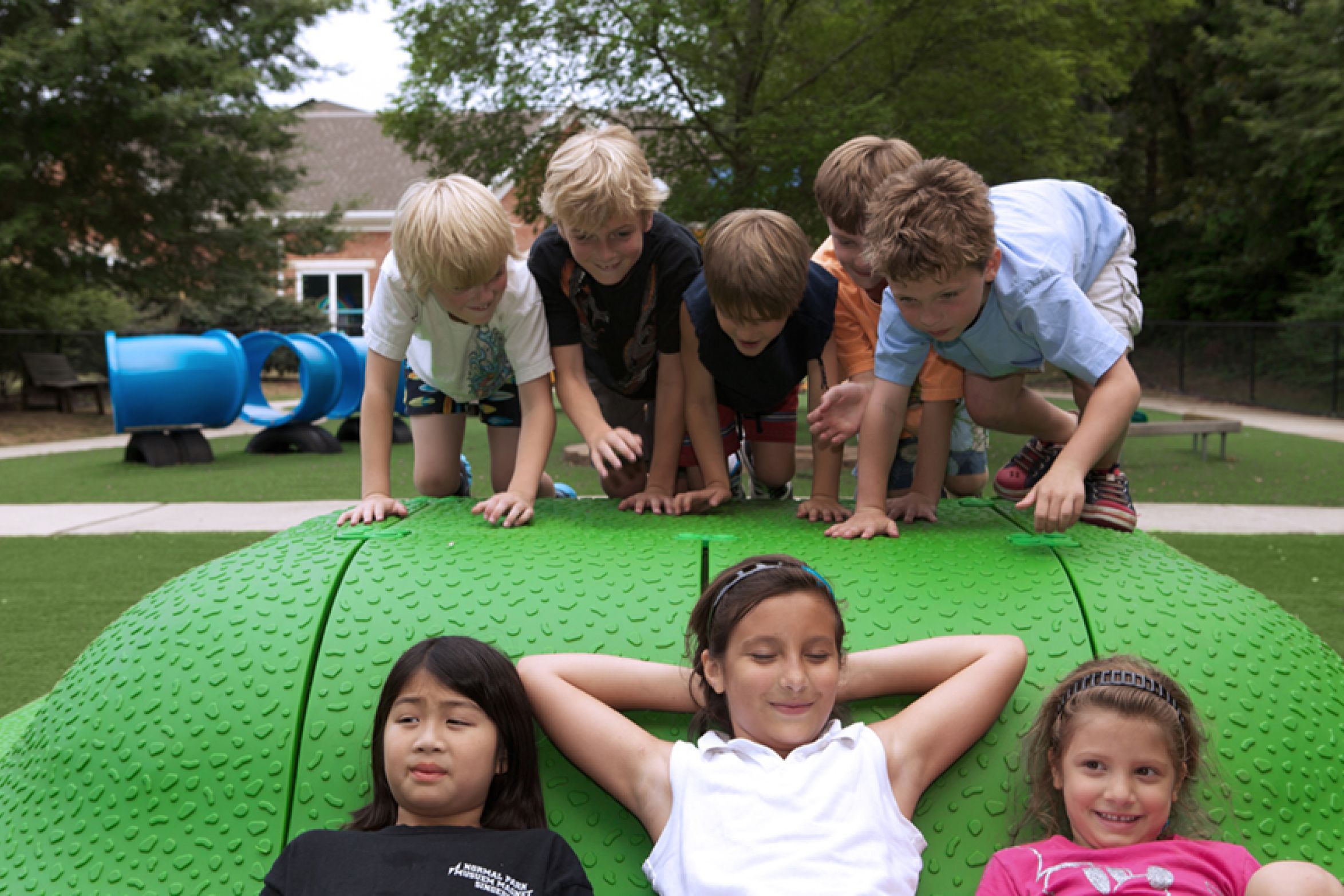 Children playing on a textured green interactive play mound with blue tunnel slides and surrounding trees