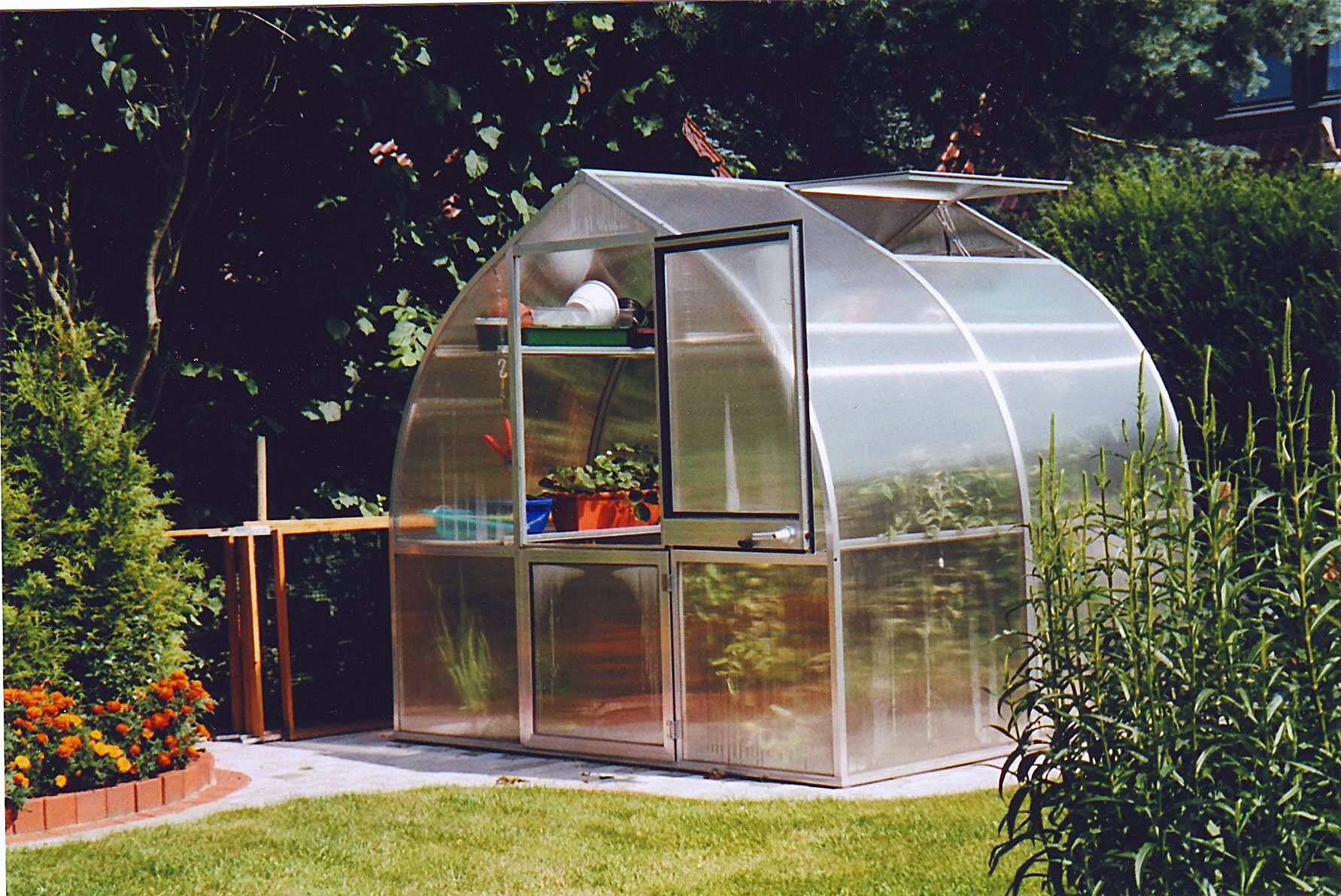 Compact greenhouse with curved twin-wall polycarbonate panels, roof vent, barn-style door, and potted plants inside.