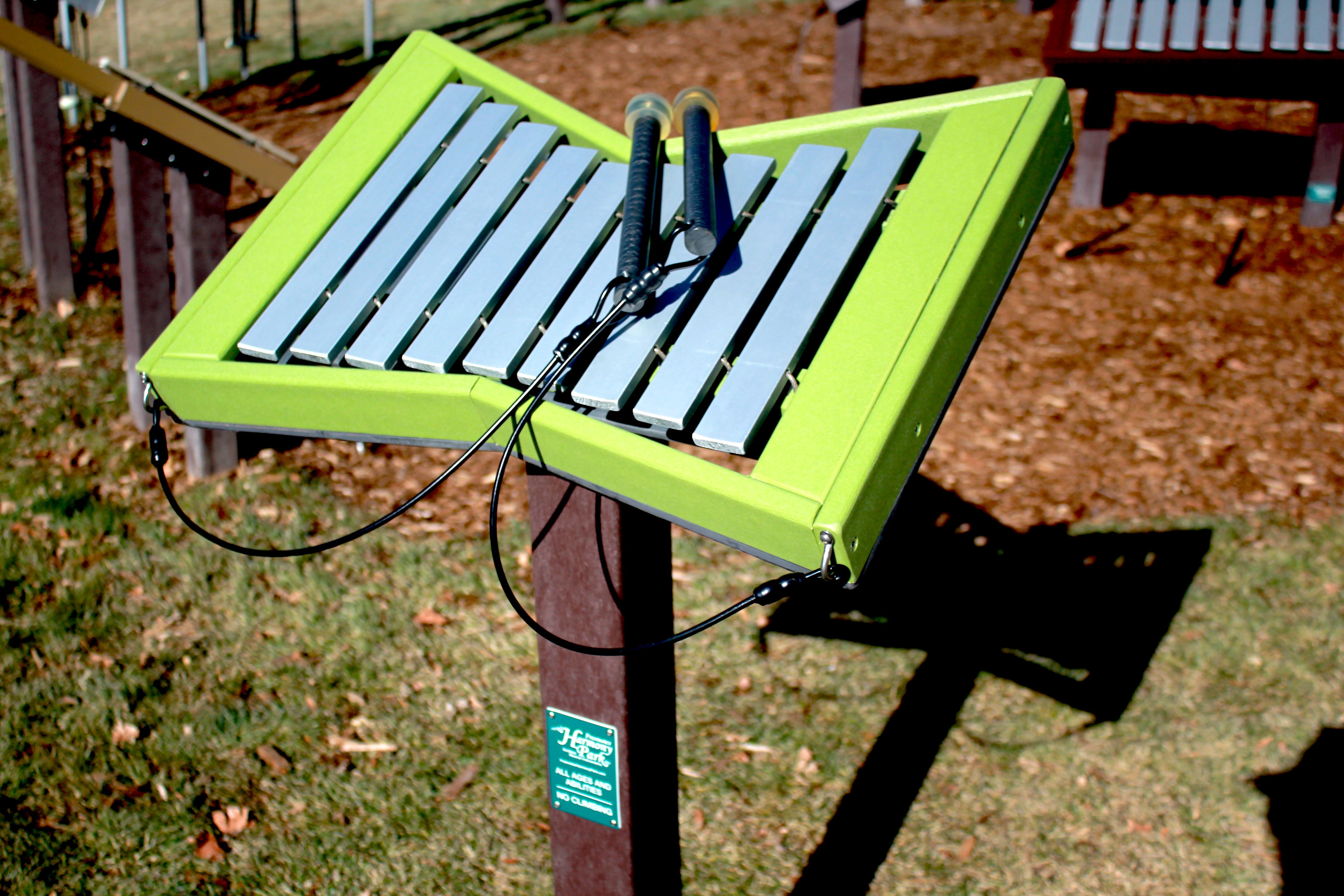 Toddler-height outdoor musical instrument with metal bars and green frame mounted on a post with attached mallets