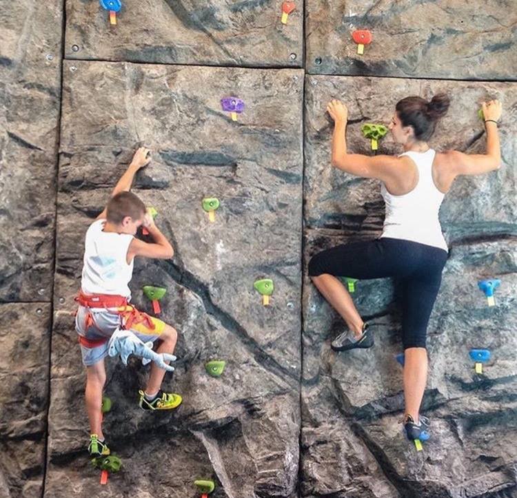 Two climbers on a modular climbing panel with colorful handholds and textured rock-like surfaces