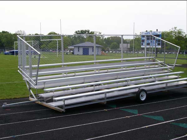 Five-row portable aluminum bleachers with chain-link guardrails and wheels on a track field.