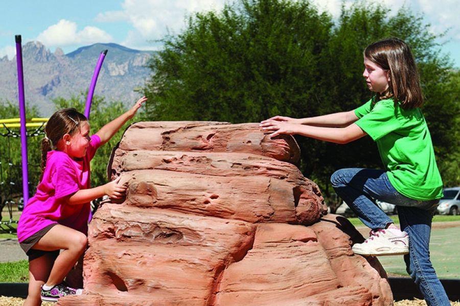 Children climbing a small textured sandstone-style climbing boulder in an outdoor playground setting