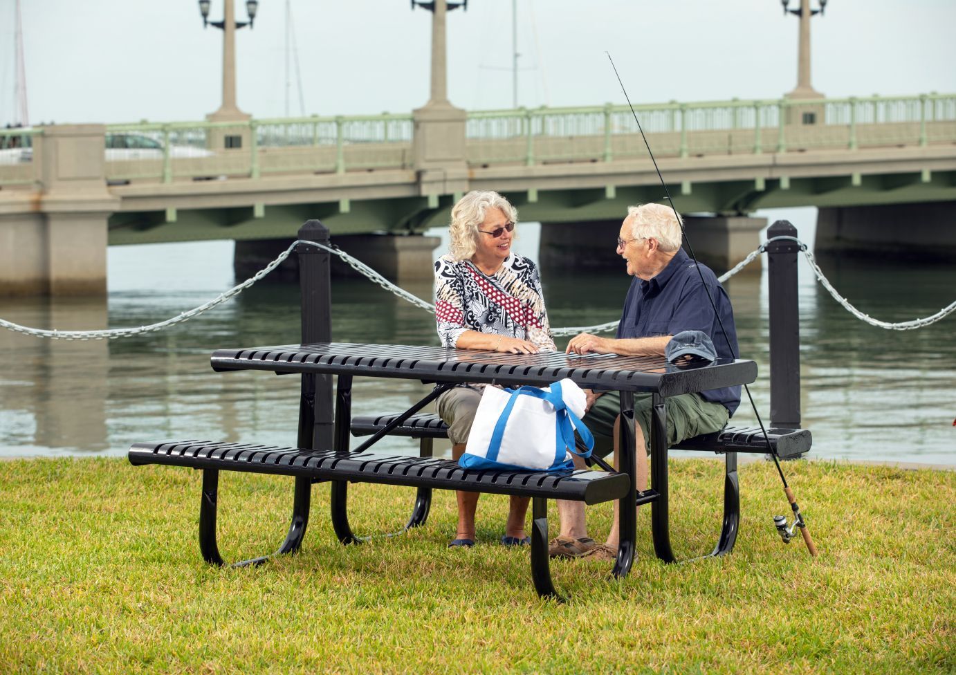 Black thermoplastic picnic table with attached benches, two elderly people seated, fishing rod and tote bag by waterfront