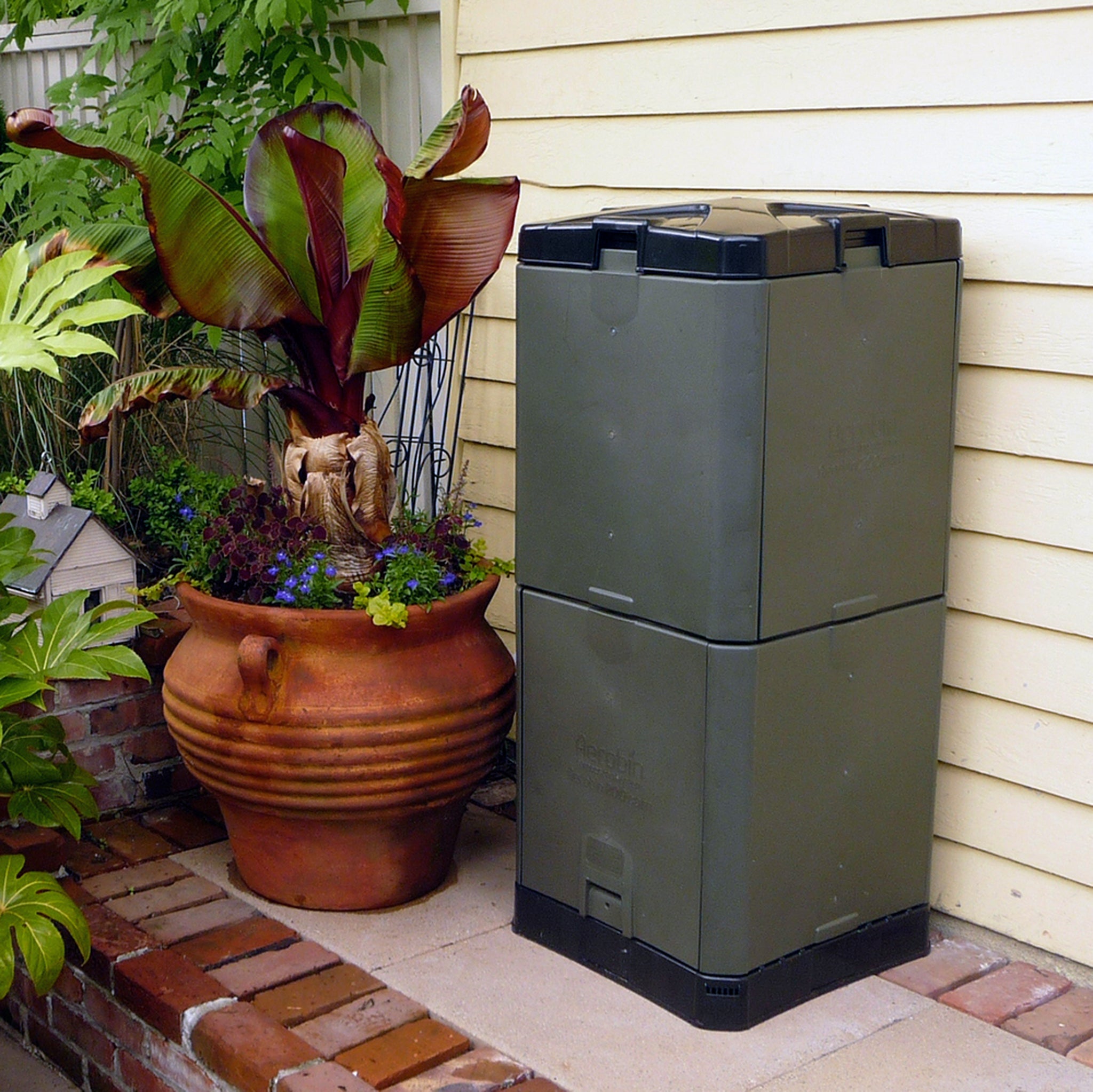 Grey insulated compost bin with black lid next to large terracotta planter with tropical plants outdoors