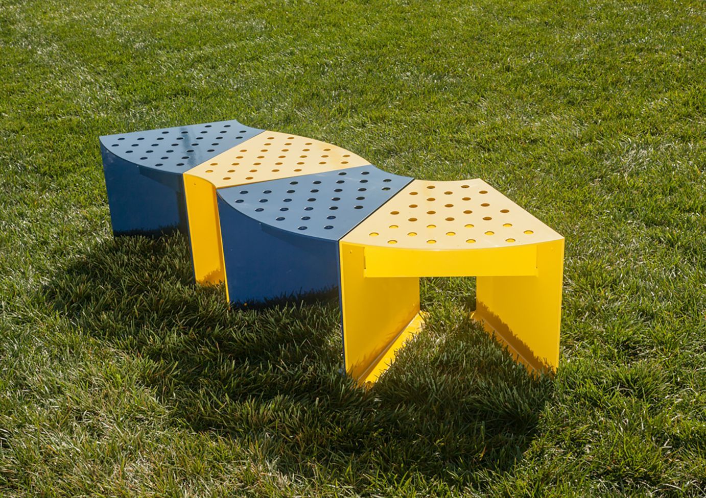 Modular outdoor bench with alternating yellow and blue perforated steel segments on grass