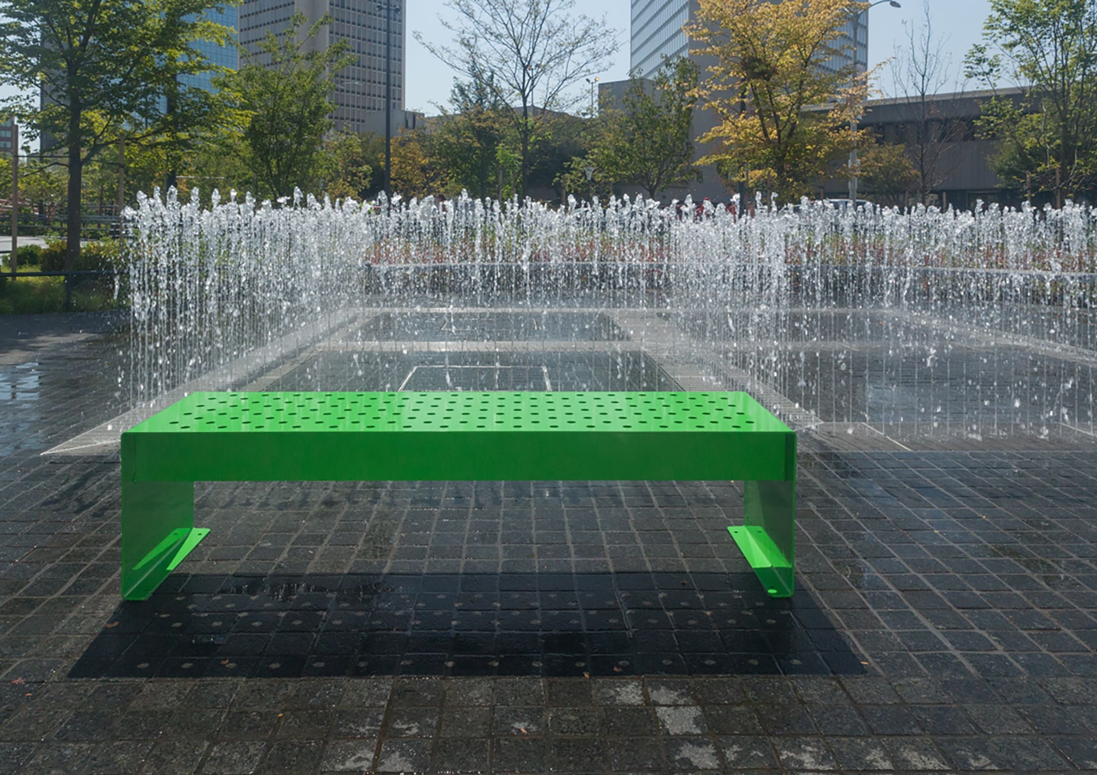 Bright green powder-coated steel bench with perforated seat, outdoor near decorative water fountain.