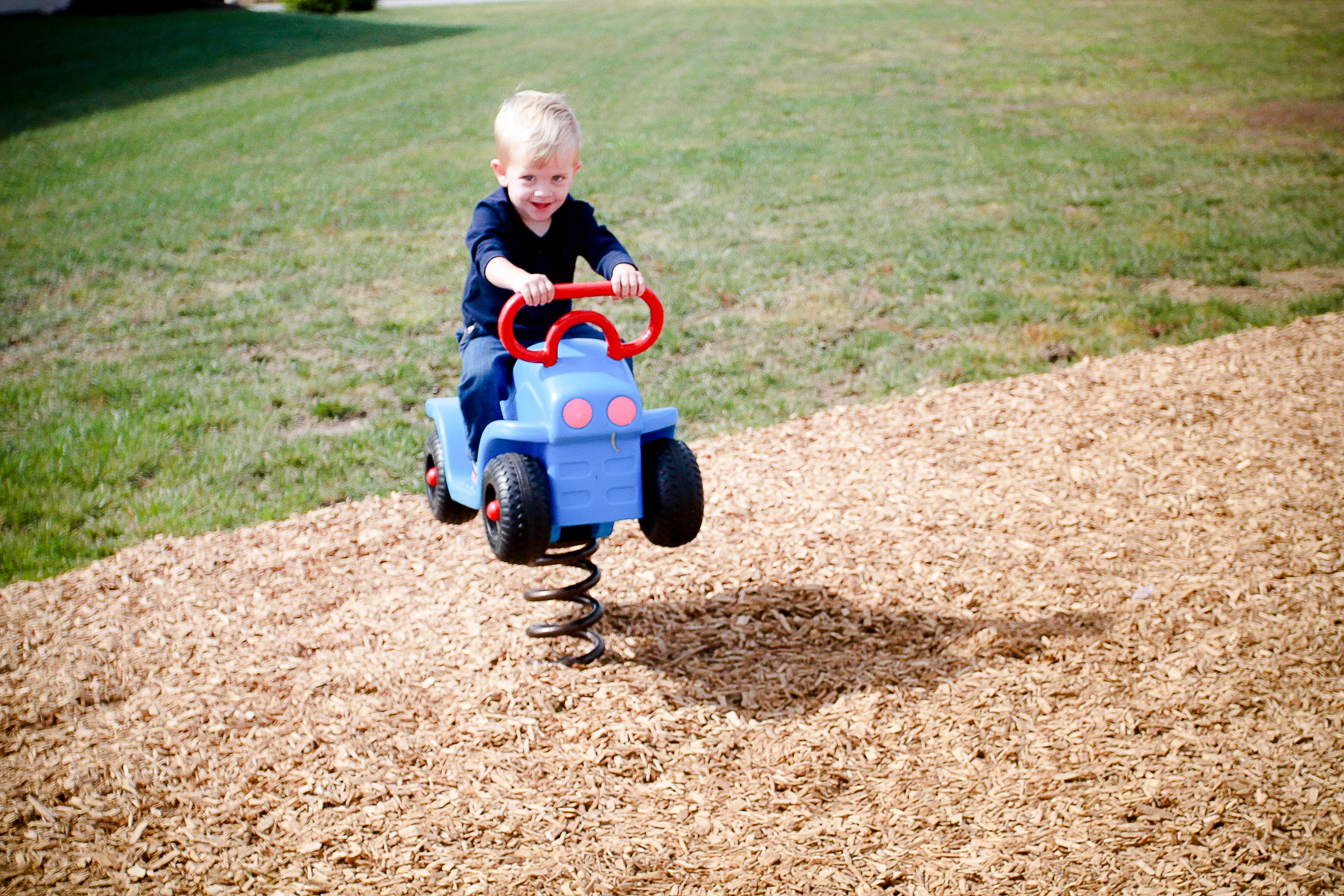 Young child on blue spring-mounted playground truck with red handle and spinning wheels on wood chip surface