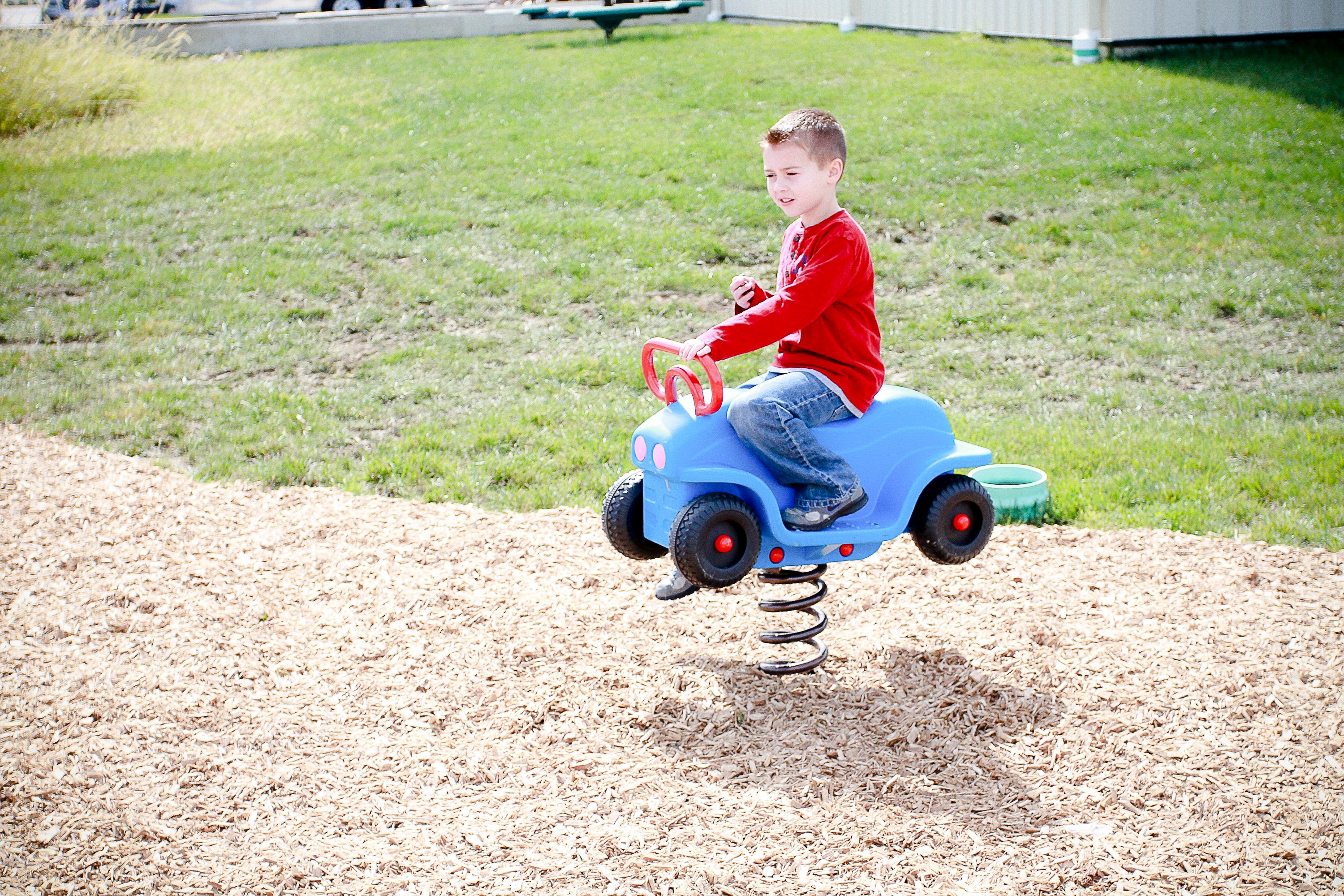 Child in red shirt on blue spring-mounted truck playground toy with spinning wheels on wood chip surface