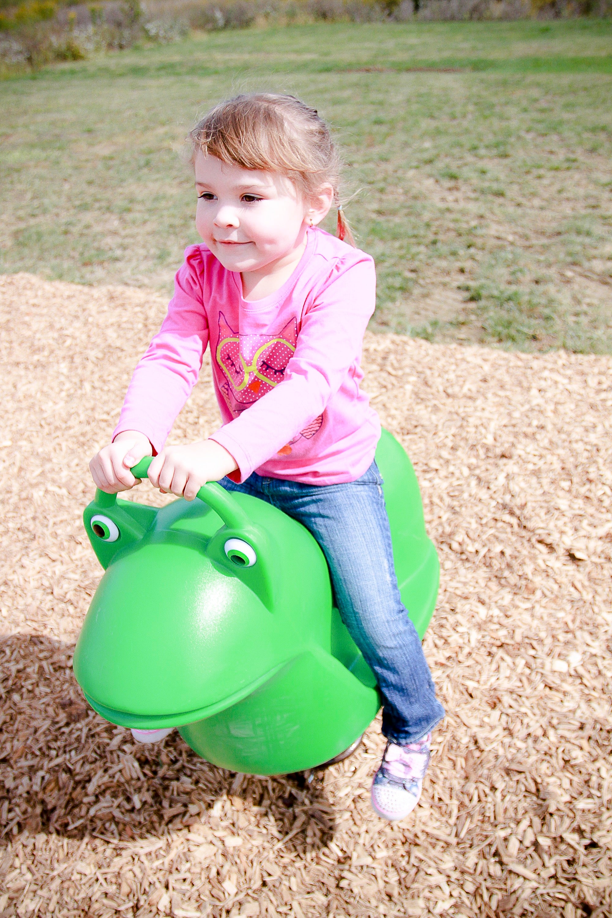 Young child on bright green frog-shaped spring rider playground toy on wood chip surface