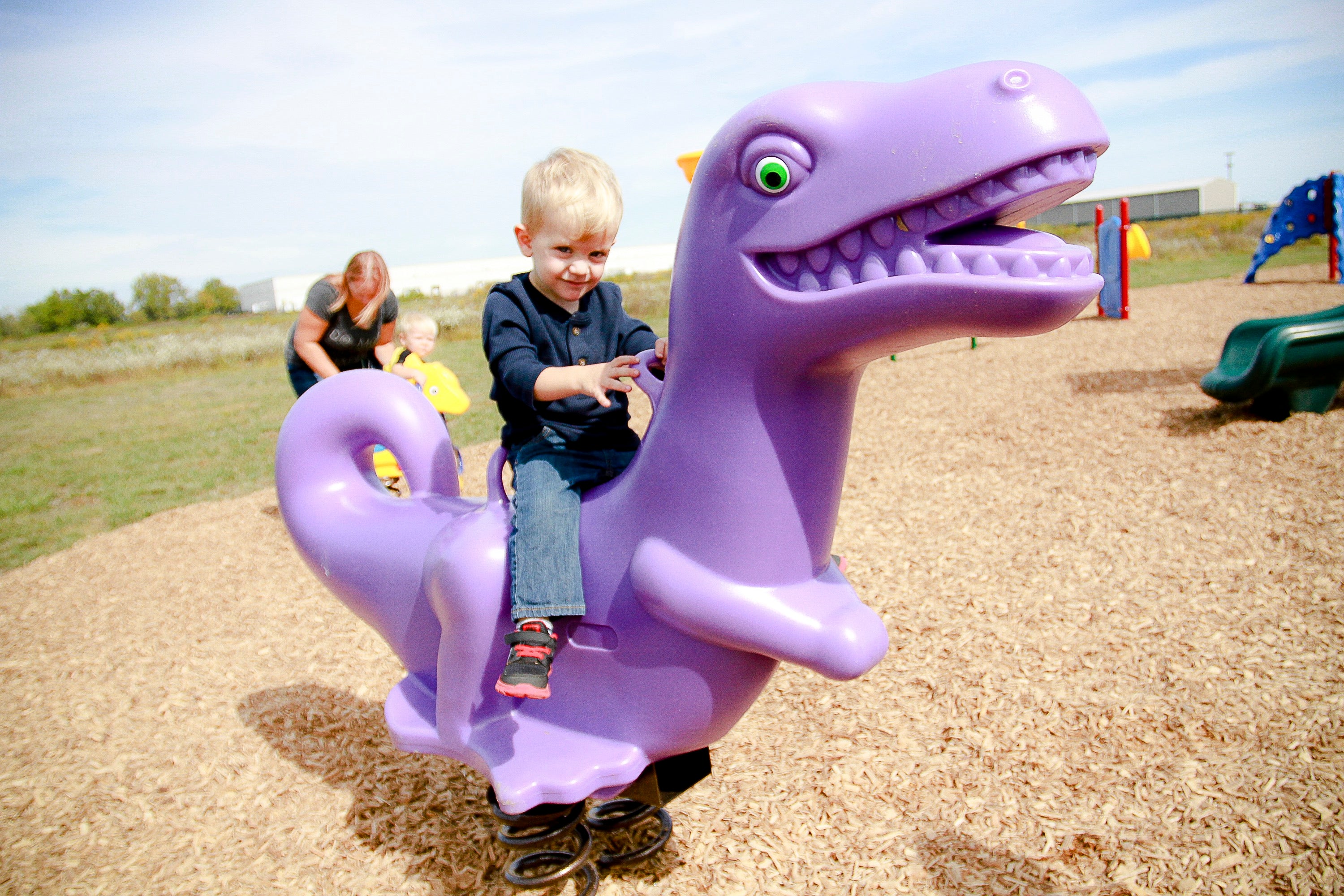 Purple dinosaur-shaped playground spring rider with molded handholds and footrests on wood chip surface with child seated