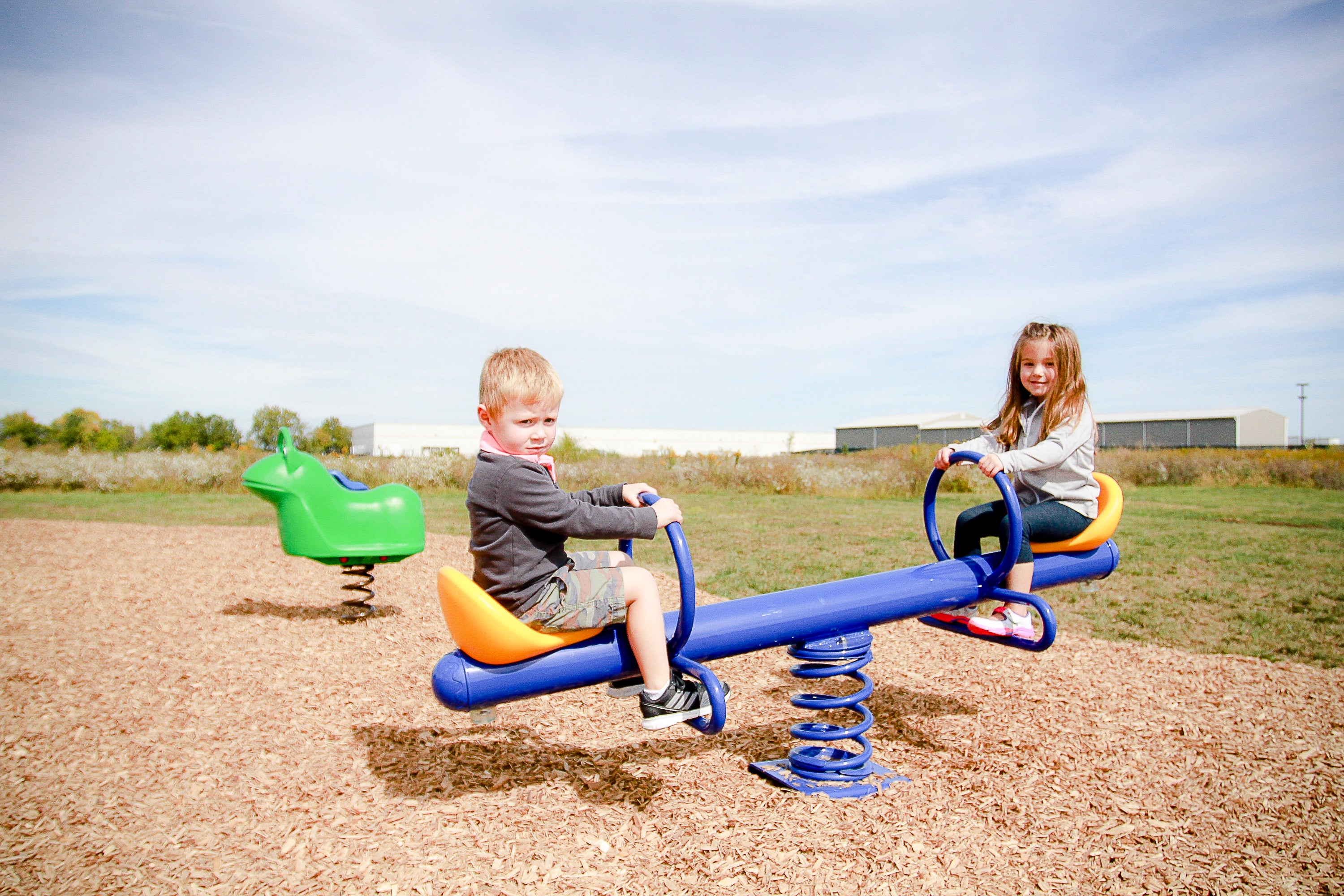 Outdoor playground spring see saw with blue beam, orange seats, molded back support, and two children playing on wood chips