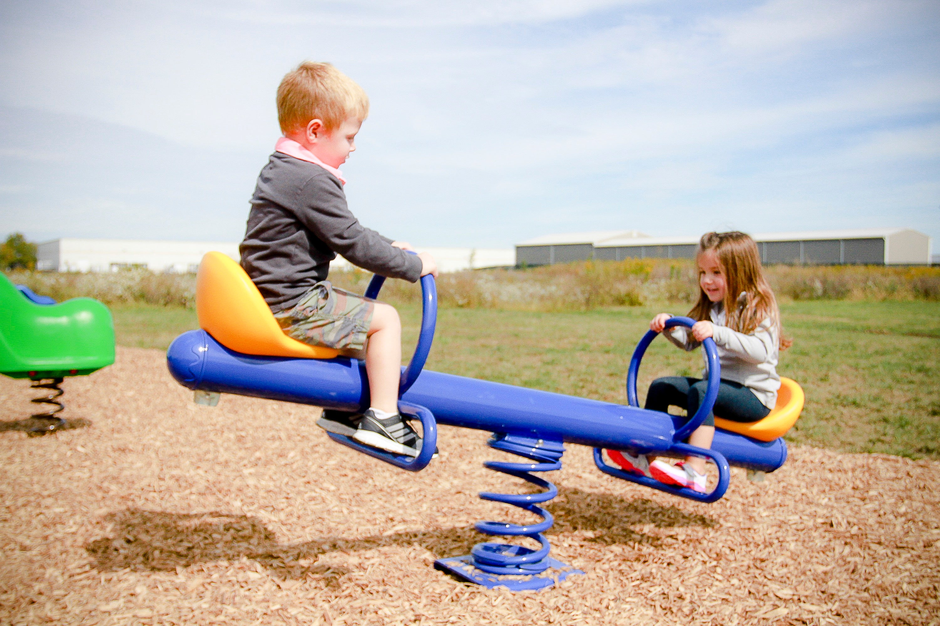 Two children on a blue spring-mounted see saw with orange molded seats and blue handles on a wood chip playground