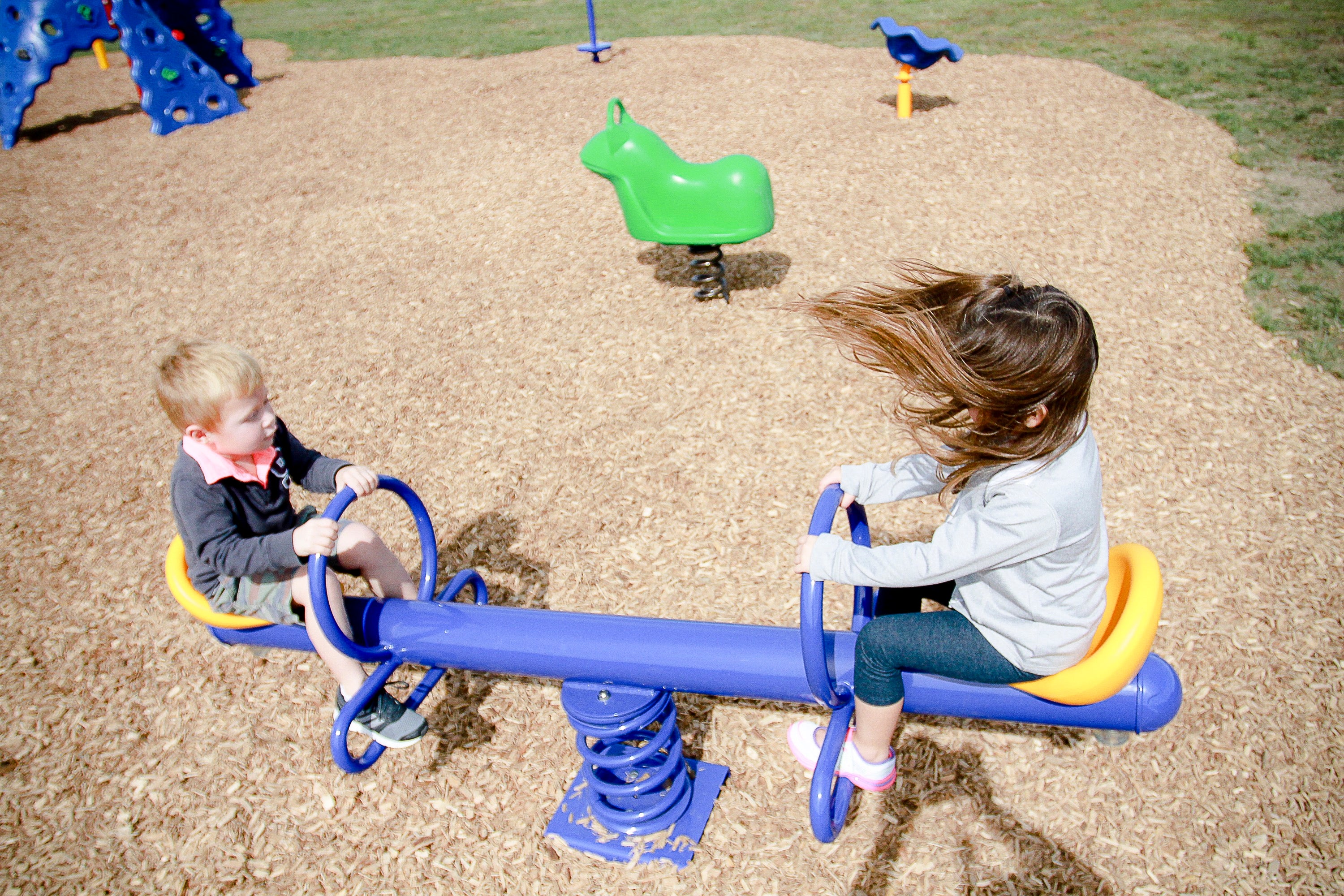 Playground see saw with blue and yellow molded seats, spring-mounted, two children playing on wood chip surface