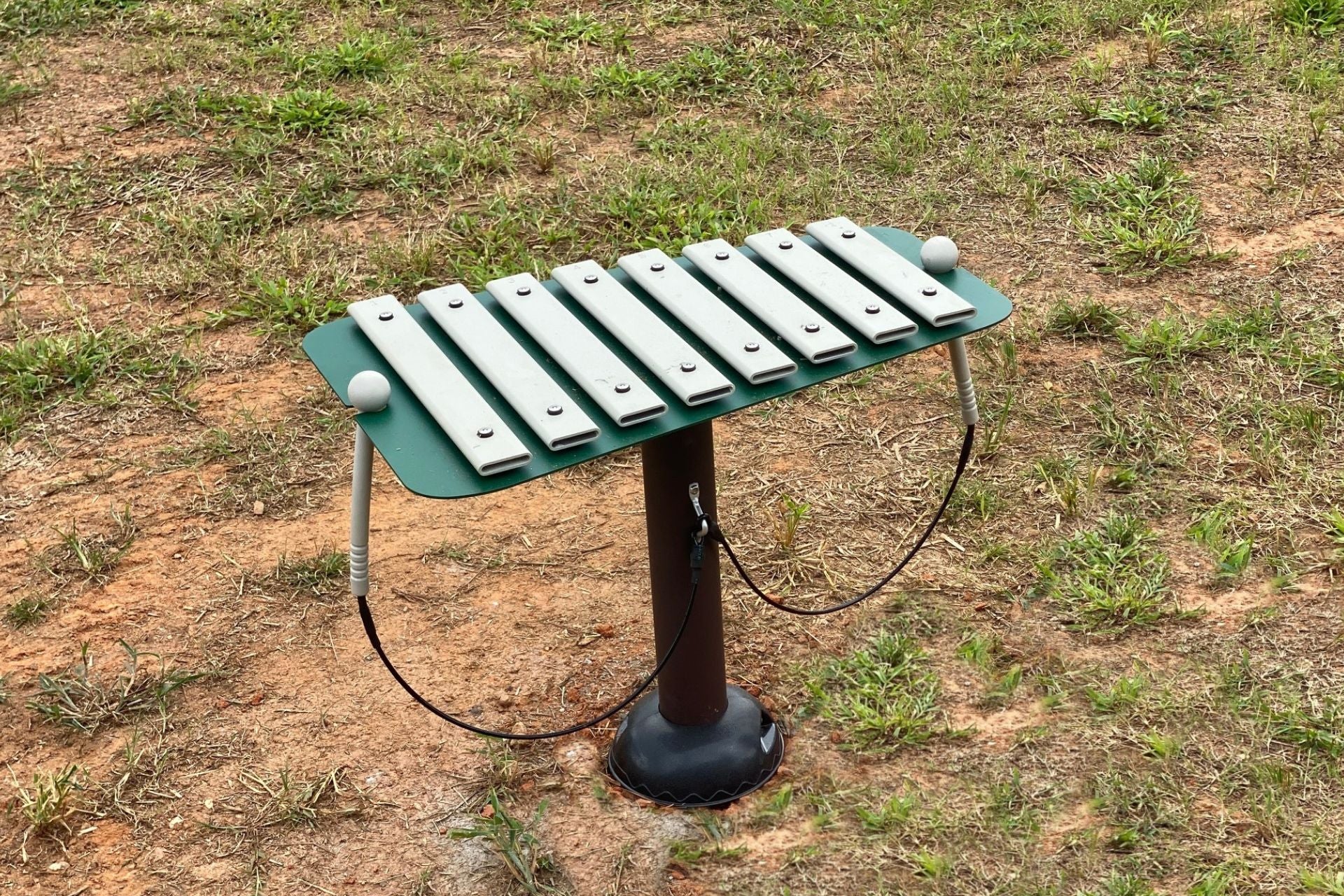 Outdoor playground glockenspiel with metal bars and attached mallets on a green frame mounted on a single post