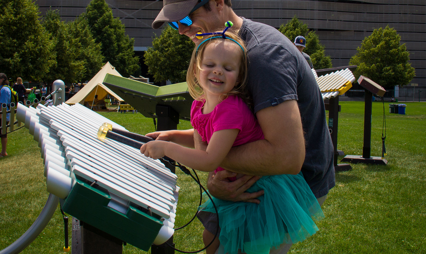 Child in colorful outfit playing silver and green outdoor metallophone on grassy park lawn with adult nearby