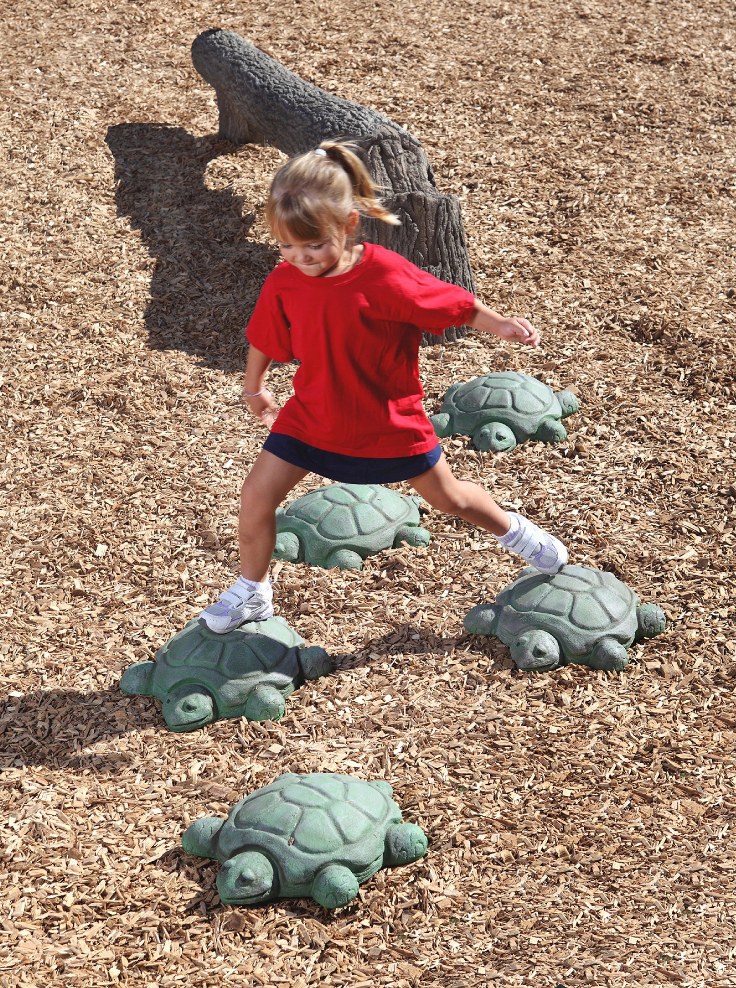A child playing on nature-themed turtle steppers in an outdoor playground setting, surrounded by wood chips and a log structure.
