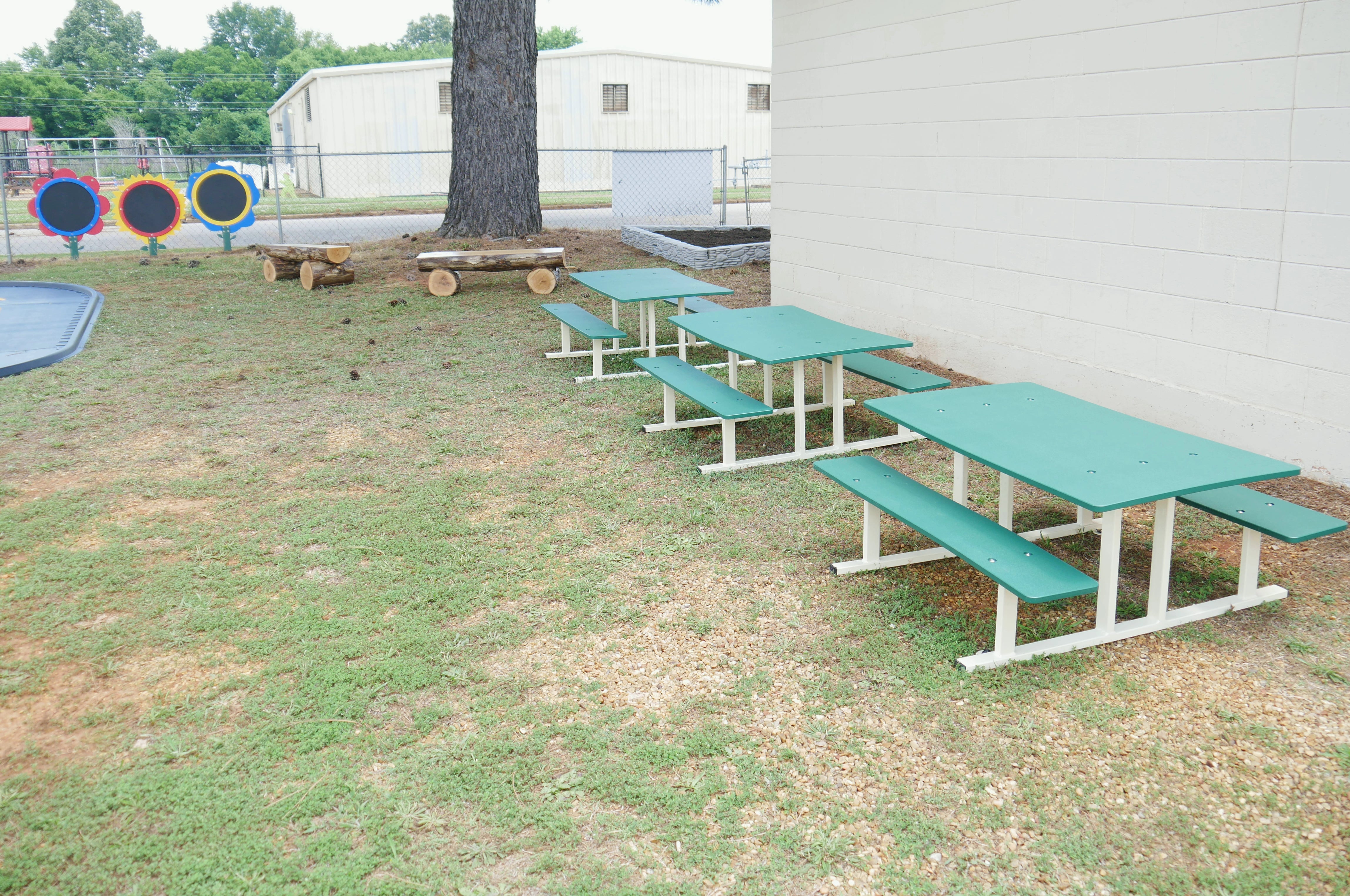 Three green preschool picnic tables with white metal frames arranged outdoors near a building wall