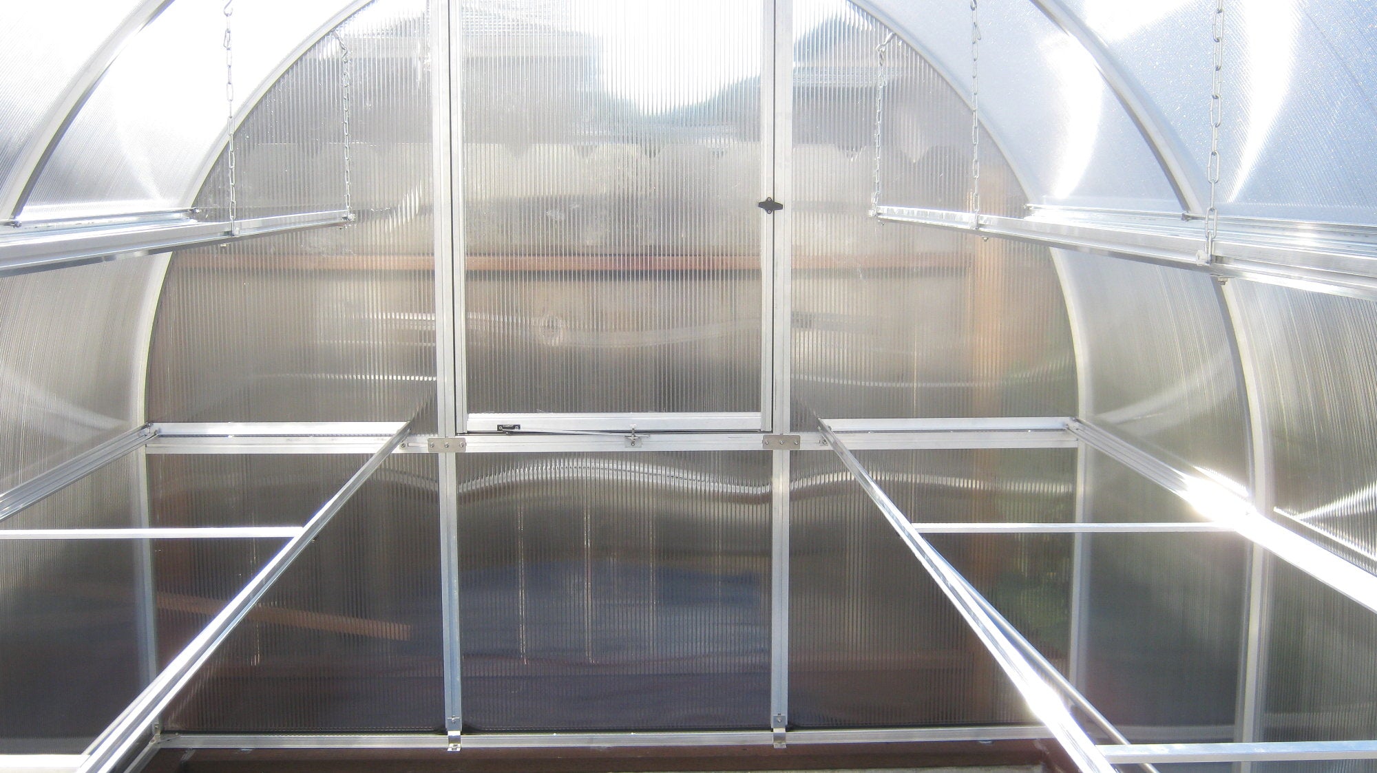 Interior of greenhouse with clear twin-wall polycarbonate panels, metal shelving, rear ventilation window, and roof vent