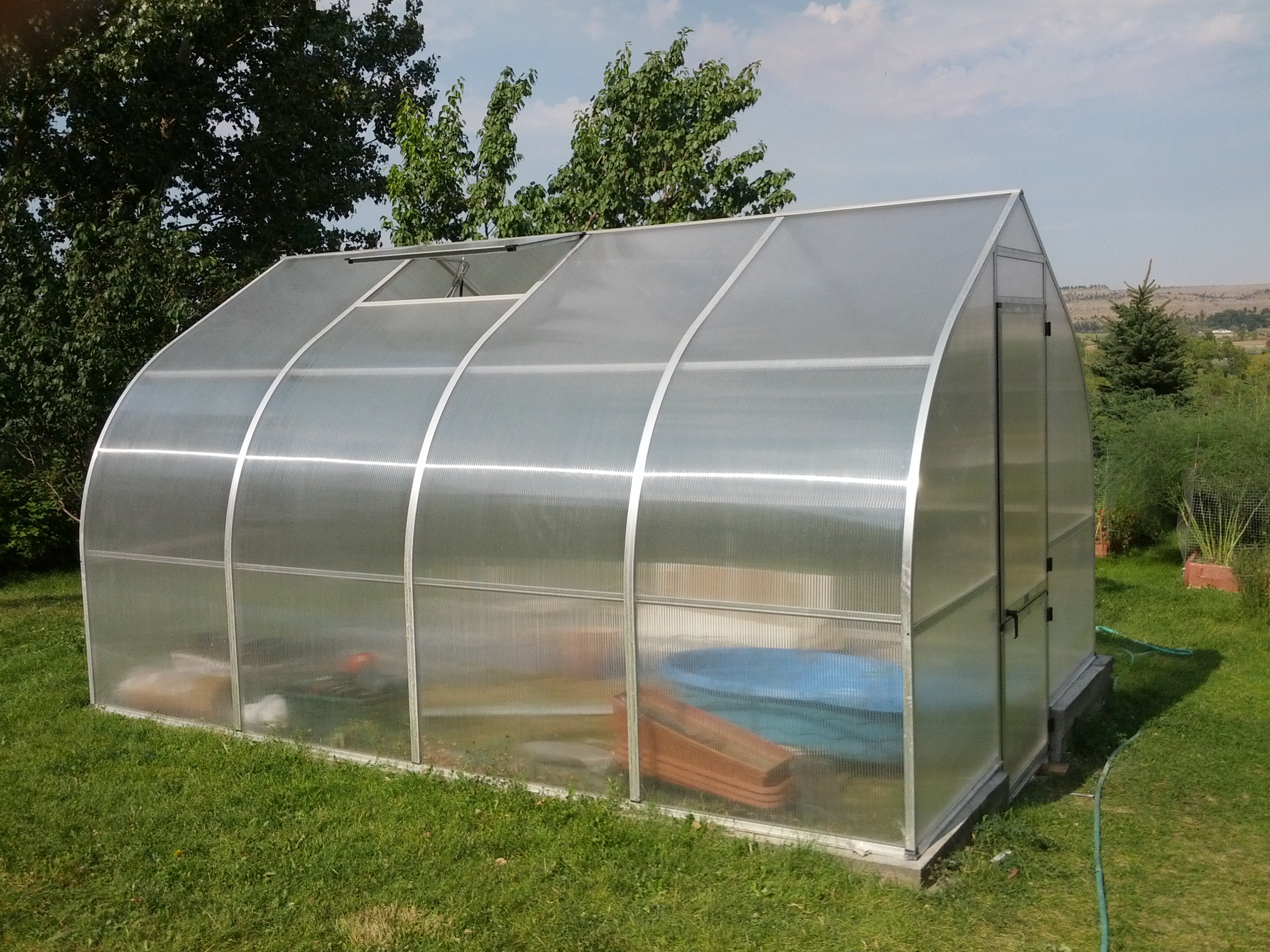 Curved greenhouse with translucent twin-wall polycarbonate panels, roof vents, barn-style door, and gardening supplies inside