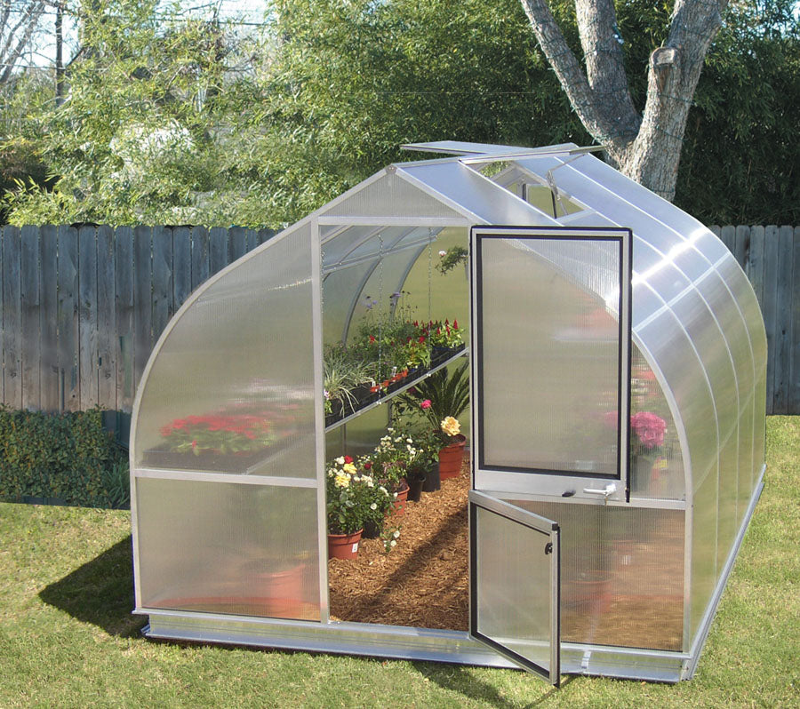 Compact greenhouse with twin-wall polycarbonate panels, barn-style door, roof vents, and interior shelves with potted flowering plants.