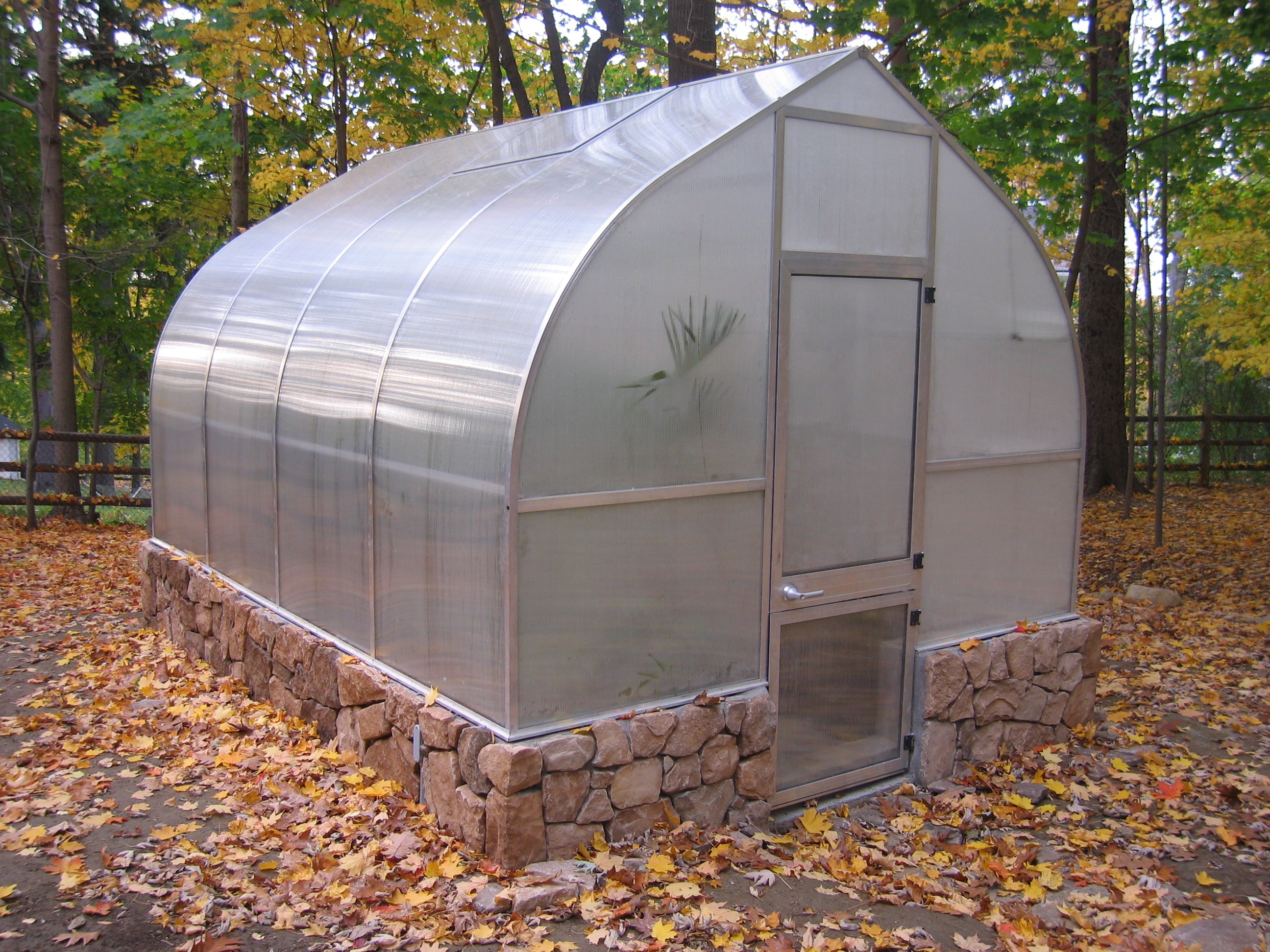 Curved twin-wall polycarbonate greenhouse with stone base, roof and rear windows, barn-style door, surrounded by autumn foliage and trees