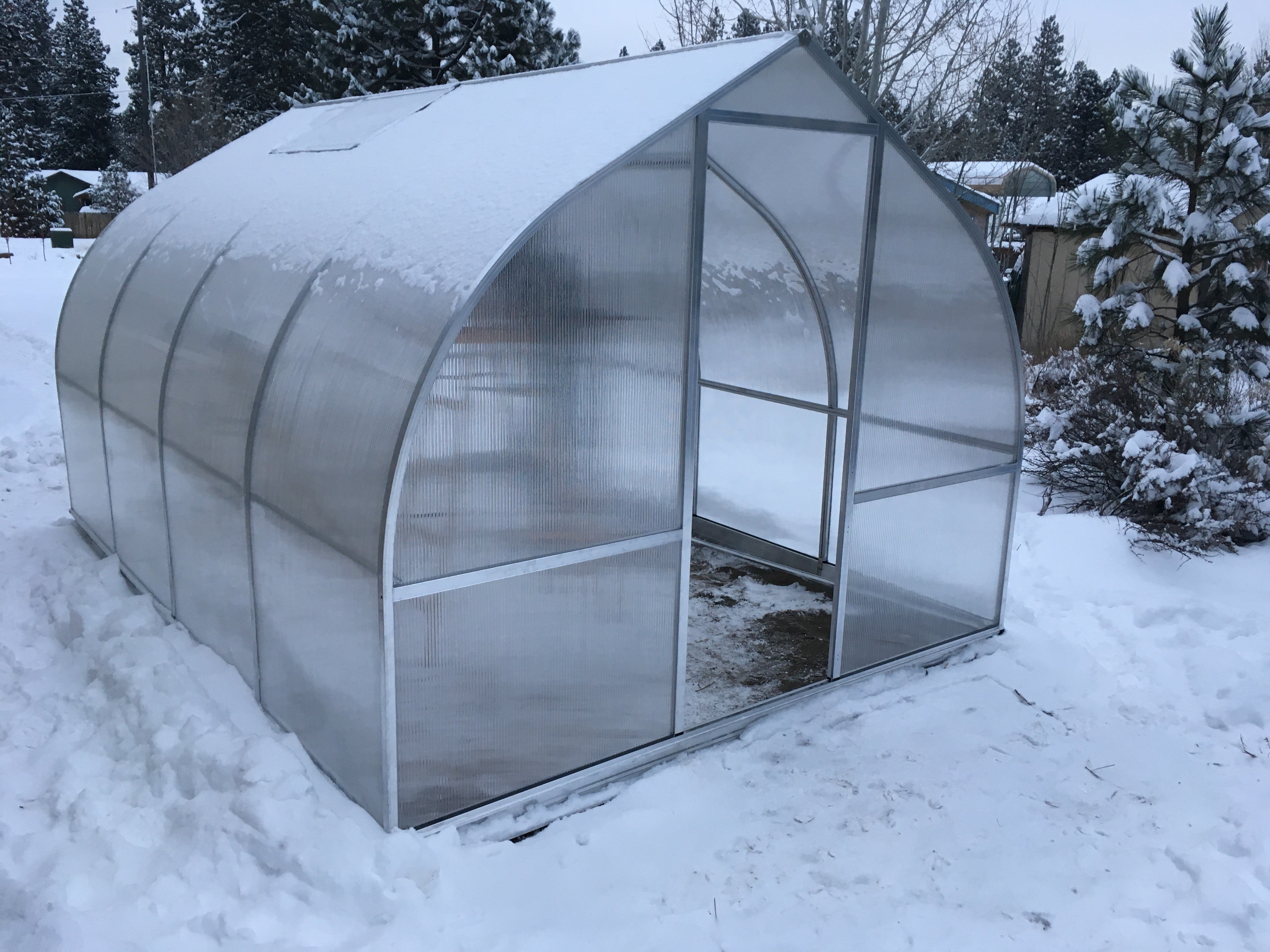 Curved polycarbonate greenhouse with barn-style door, roof vents, and snow-covered ground surrounded by trees