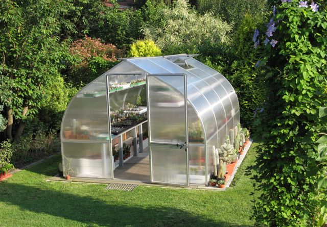 Curved polycarbonate greenhouse with automatic roof vents, barn-style door, and plants inside and outside on grass lawn