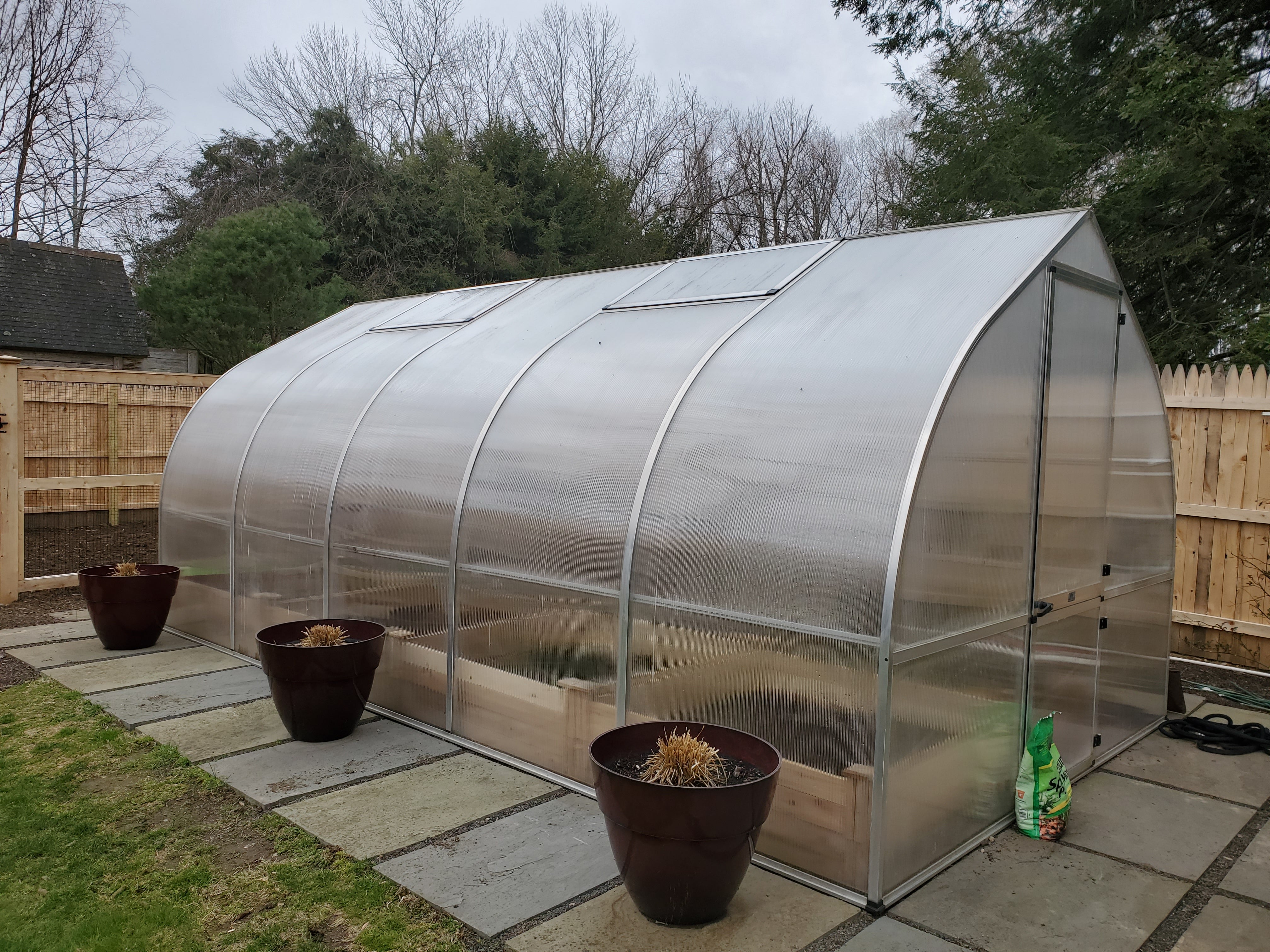 Curved polycarbonate greenhouse with automatic roof vents, barn-style door, and potted plants on stone patio