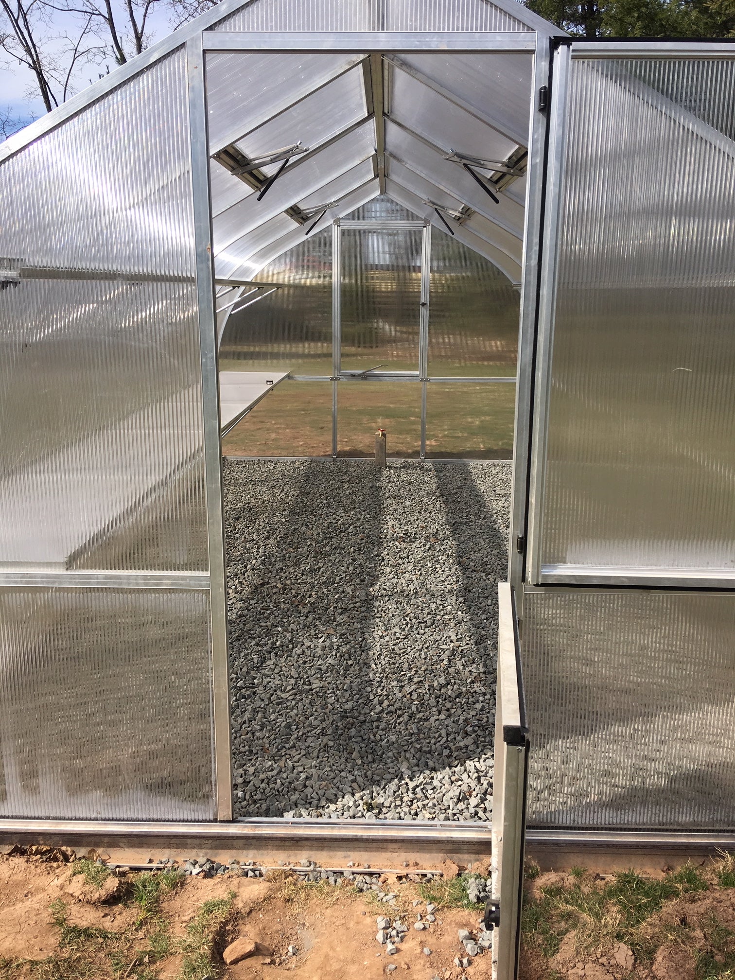 Interior of greenhouse with clear twin-wall polycarbonate panels, automatic roof vents, barn-style door, and gravel floor.
