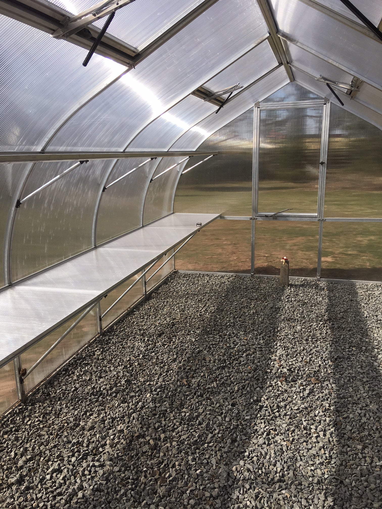 Polycarbonate greenhouse interior with metal frame, gravel floor, roof vents, barn-style door, and multiple windows