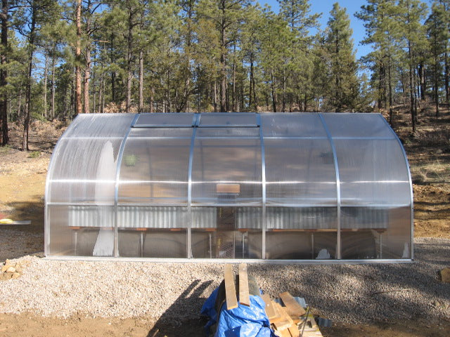 Transparent curved greenhouse with twin-wall polycarbonate panels, roof vents, barn-style door, and interior planting tables