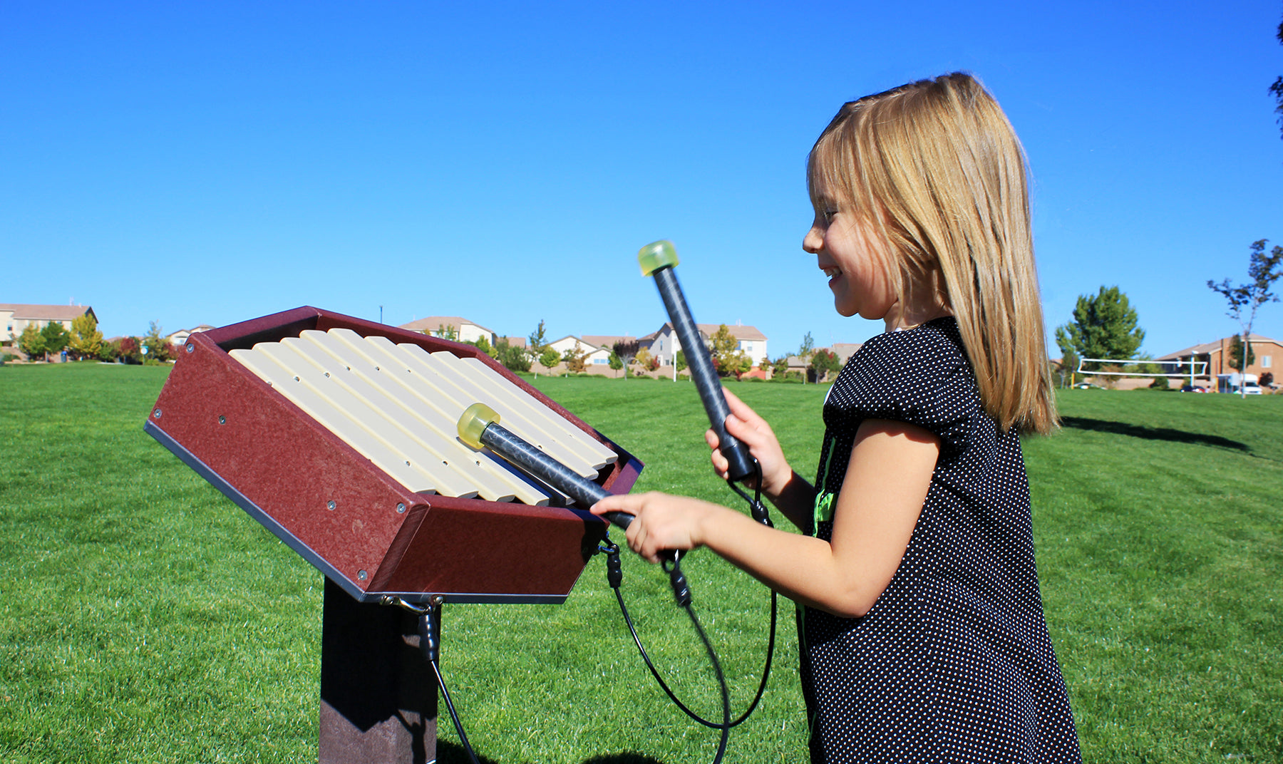 Toddler playing a toddler-height outdoor musical percussion instrument with mallets on green lawn under clear sky