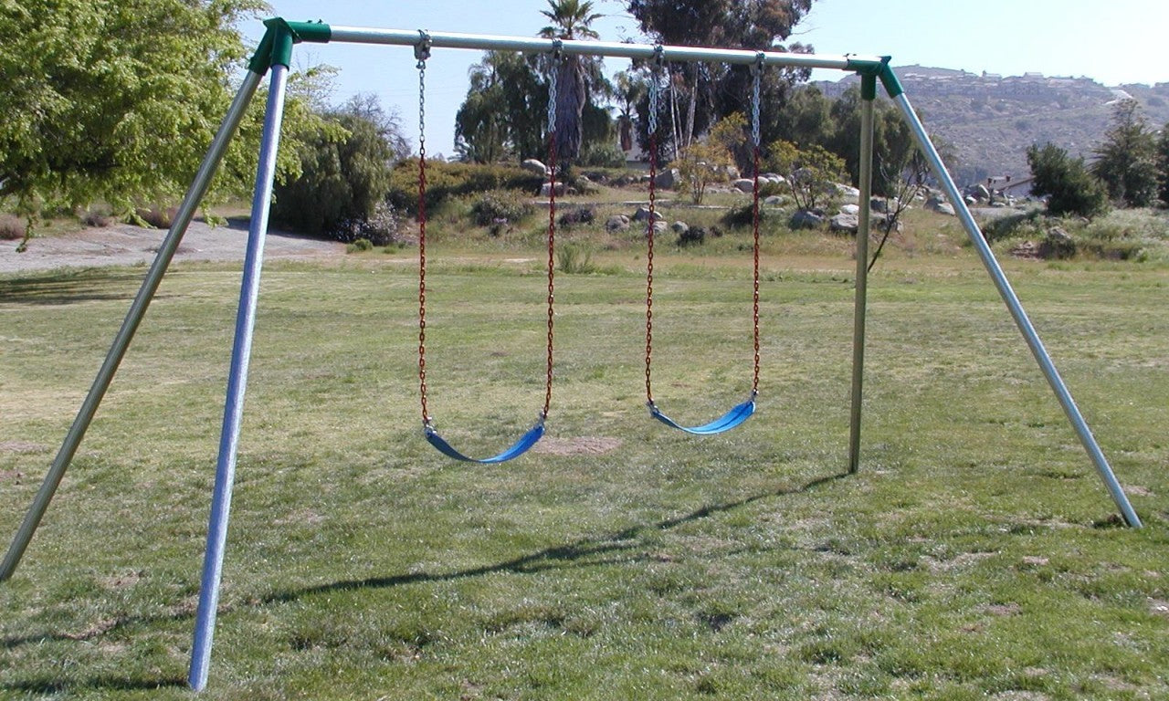 Metal swing set with two blue plastic seats and red chains on grassy field with trees and hills in background