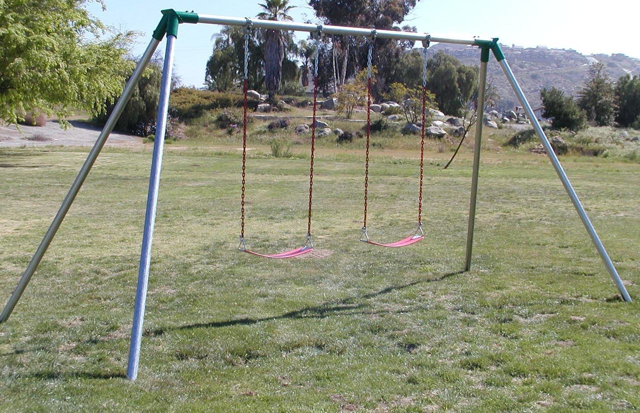 Metal swing set with two red plastic seats and chains on grassy playground, sturdy commercial play equipment