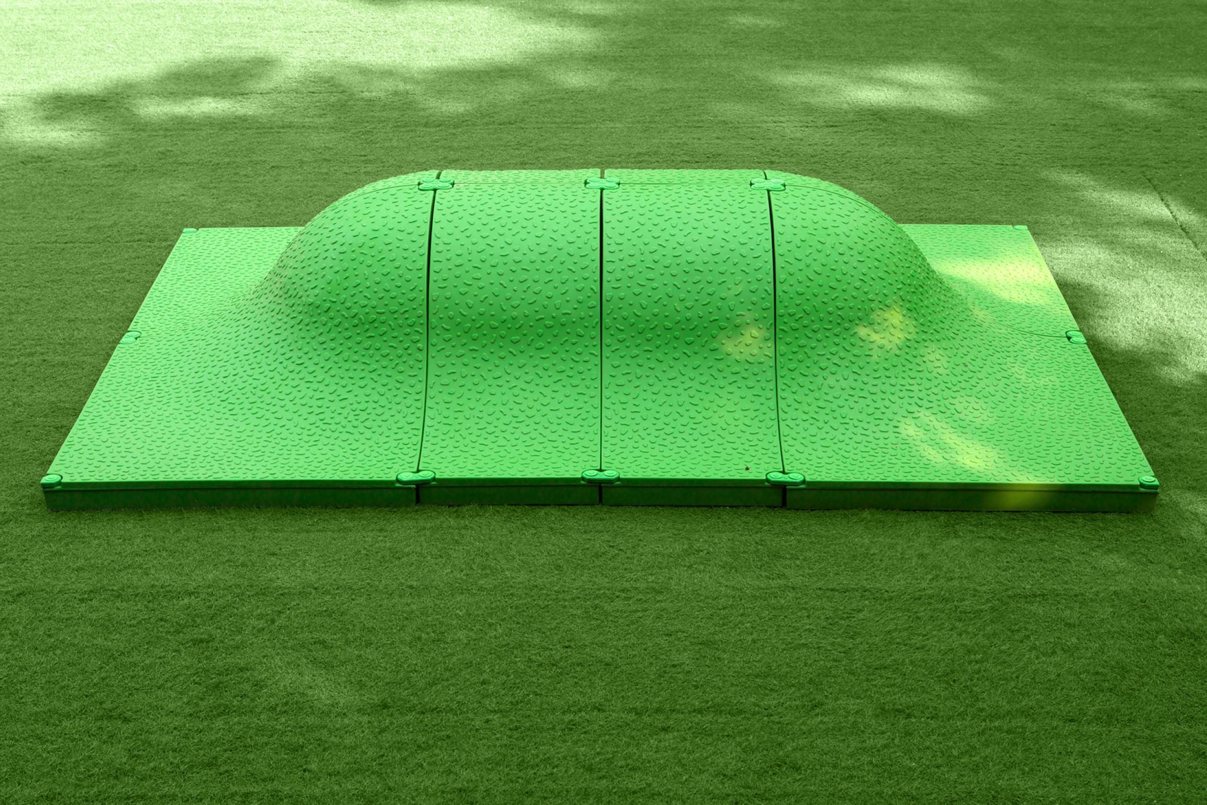 Green textured play mound with smooth curved top and segmented panels on artificial turf