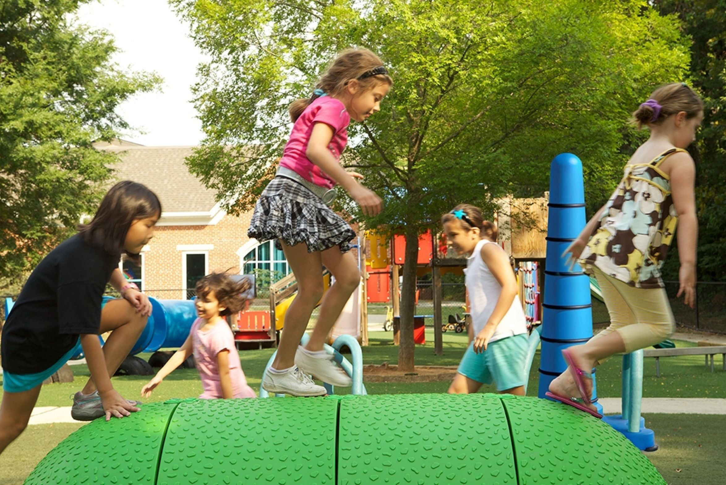 Children playing actively on a textured green interactive playground mound with colorful play equipment and trees nearby