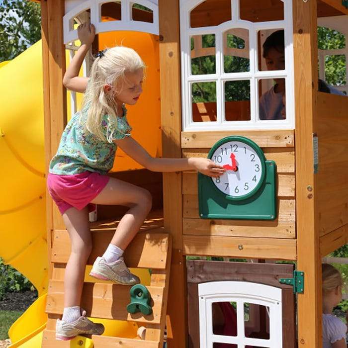 Child on wooden playset with multicolored climbing rock wall, white-framed windows, play clock, and yellow twist slide