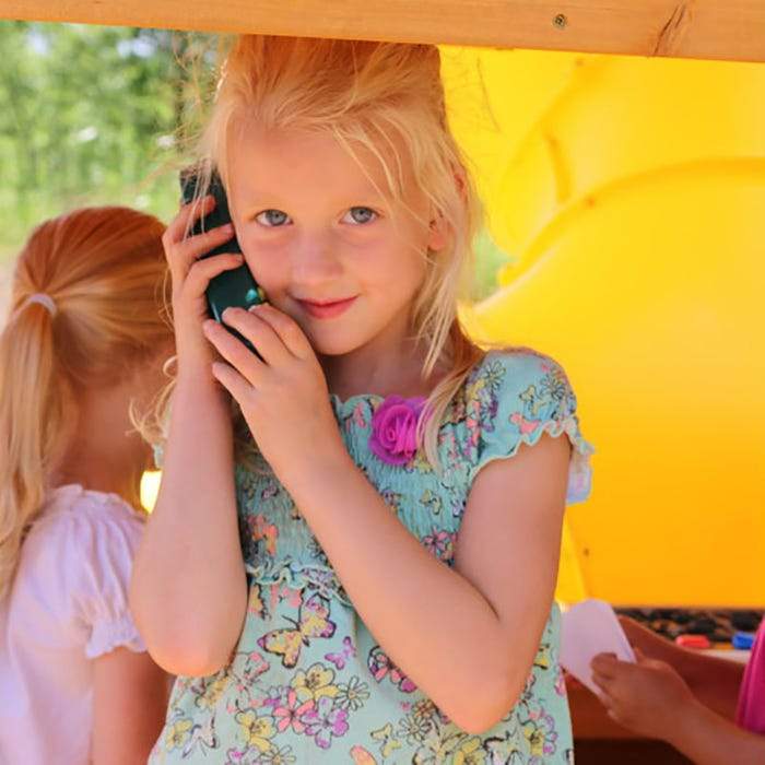 Child playing with toy phone inside wooden playset featuring bright yellow slide and multicolored climbing rockwall