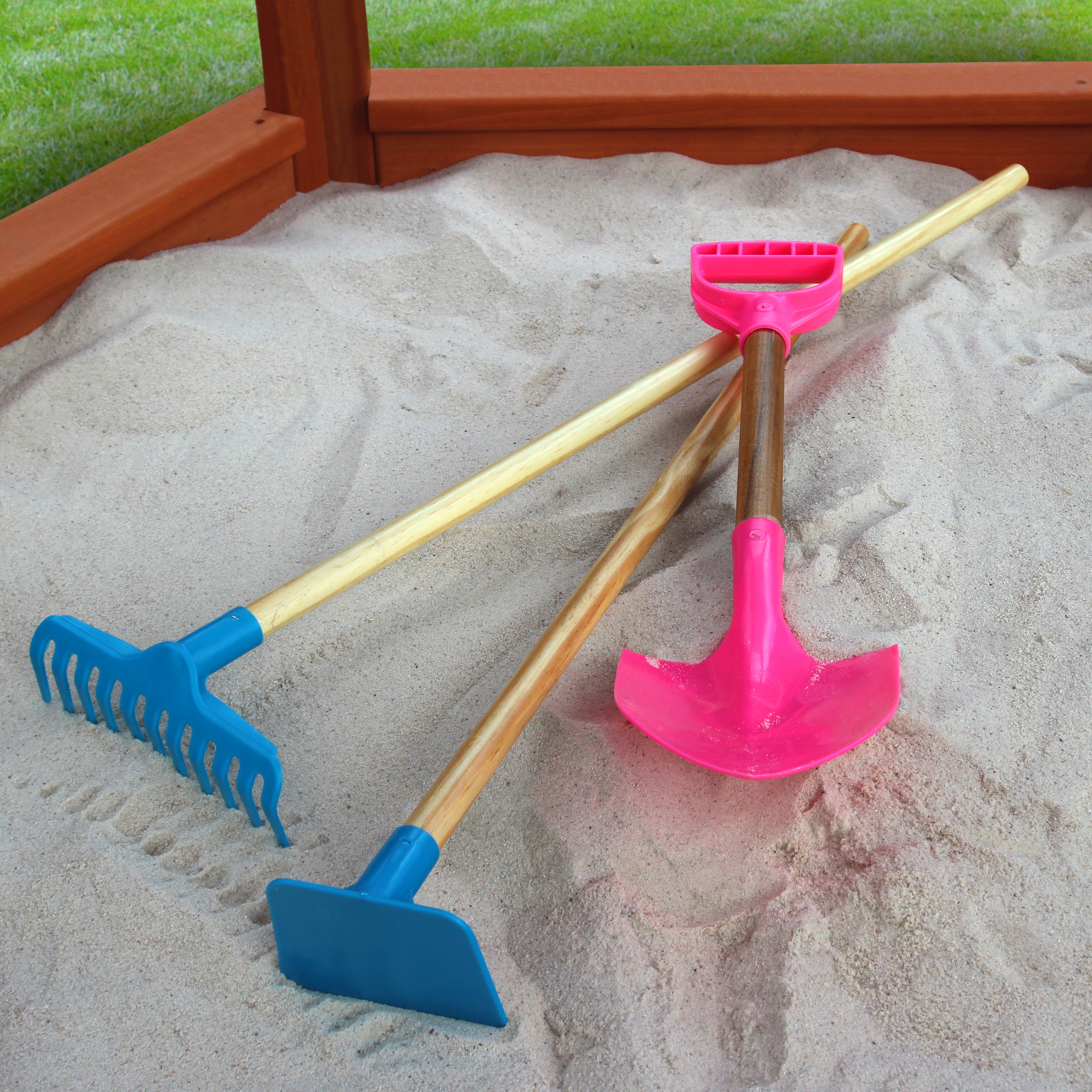 Three colorful sandbox tools with wooden handles—a pink shovel, blue rake, and blue hoe—resting on sand inside a wooden sandbox.