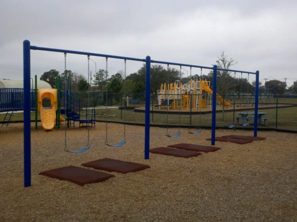 Playground swing set with blue metal frame and four swings over brown rubber safety mats on loose fill surfacing