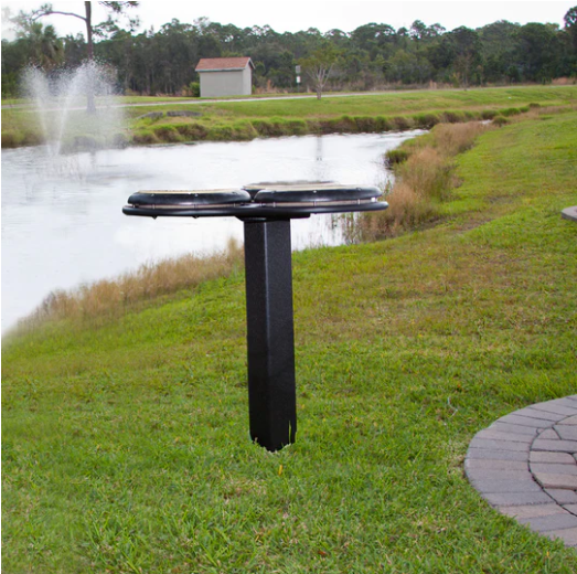 Outdoor musical drum table with three black drum pads on a single post near a pond and grassy area