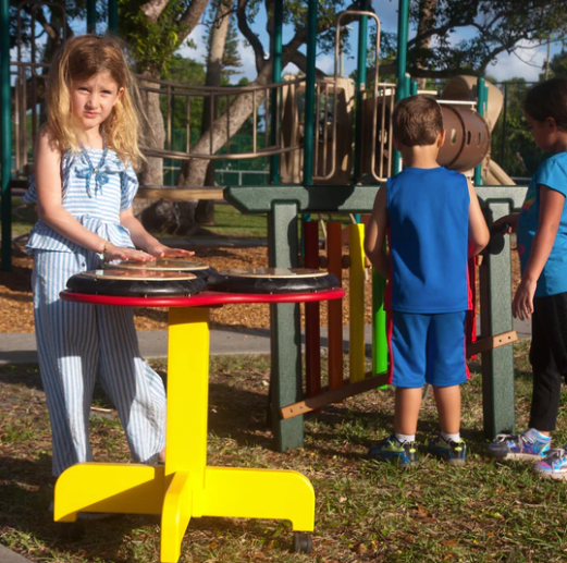 Outdoor musical drum table with three colorful drum pads and children playing in a playground setting