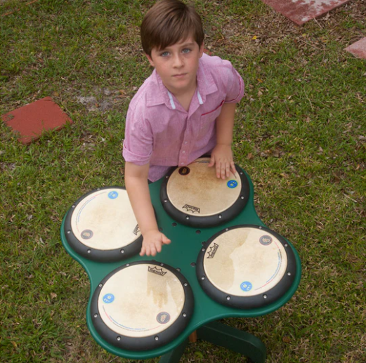 Child playing on green outdoor drum table with four professional drum heads on grass surface