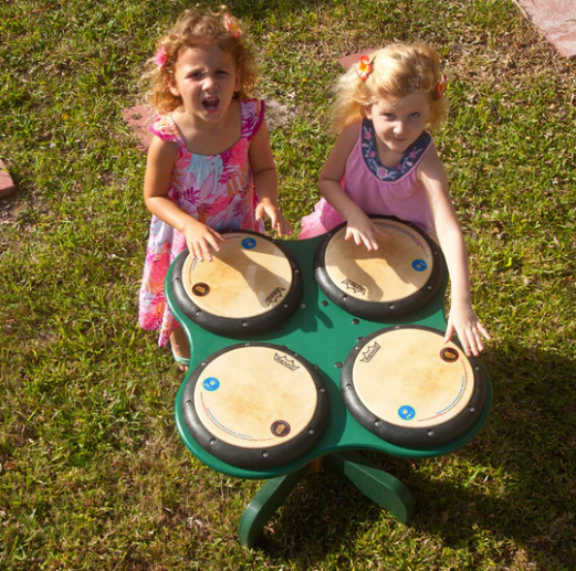 Overhead view of two children playing on a green outdoor drum table with four professional drum heads