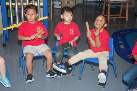 Three children seated on blue chairs playing wooden percussion claves on a colorful classroom rug with vibrant furniture