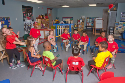 Children seated in a circle in a classroom playing colorful recycled plastic percussion instruments including tone blocks, guiros, and claves